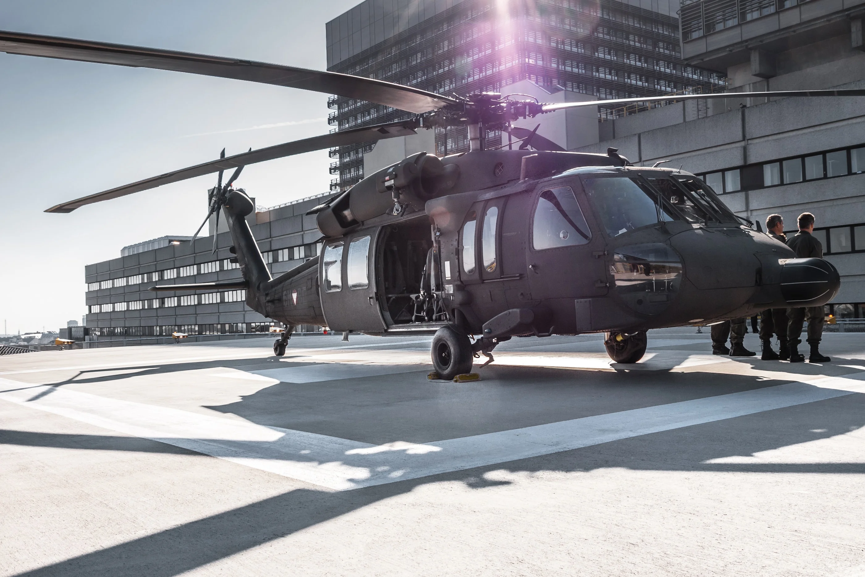 Crew beside the Black Hawk on the helipad, the hospital tower behind and the sun flaring over the rotor