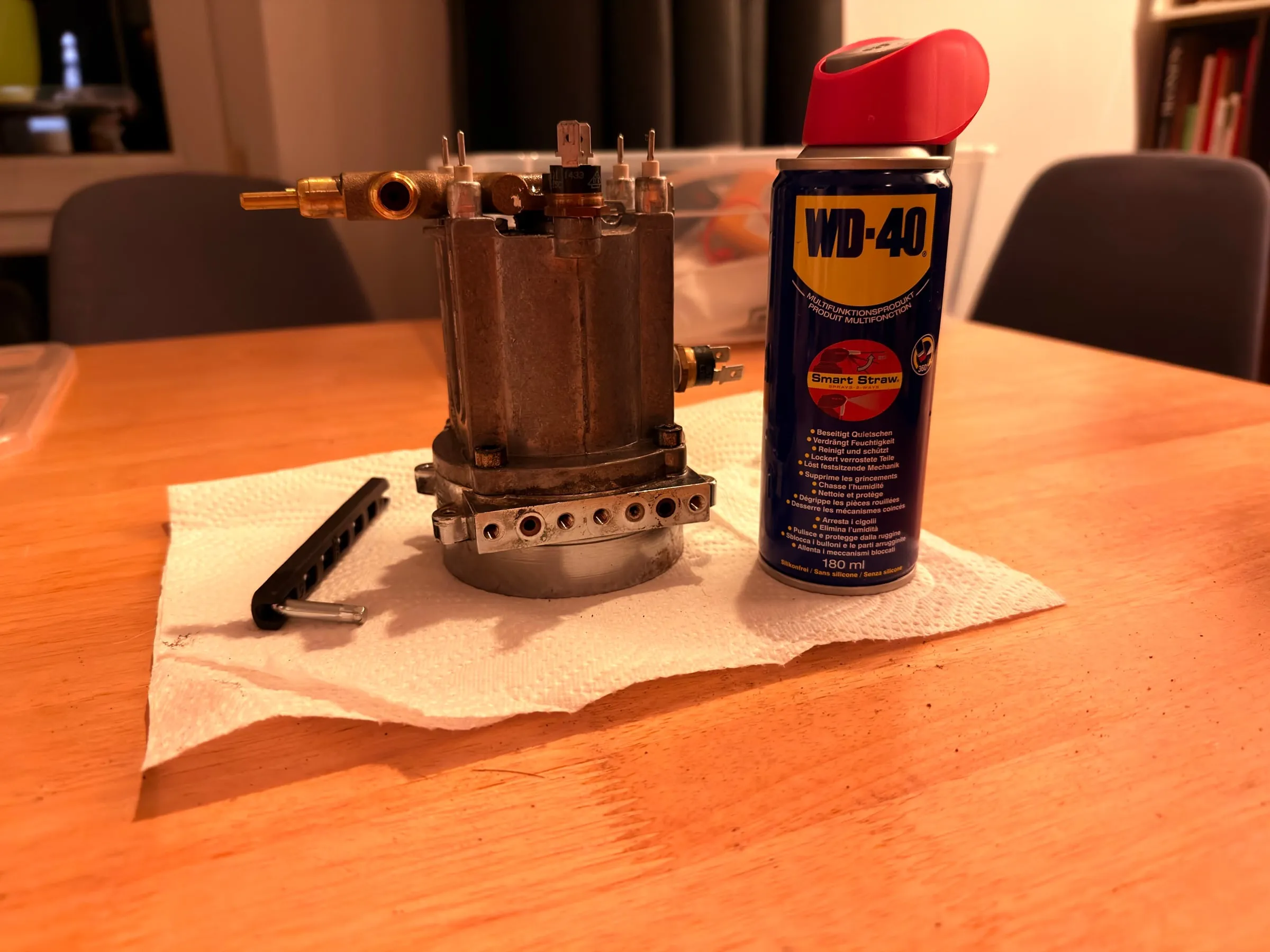 The boiler standing on kitchen paper on a wooden table at night, a can of penetrating oil beside it and a black hex key lying in front