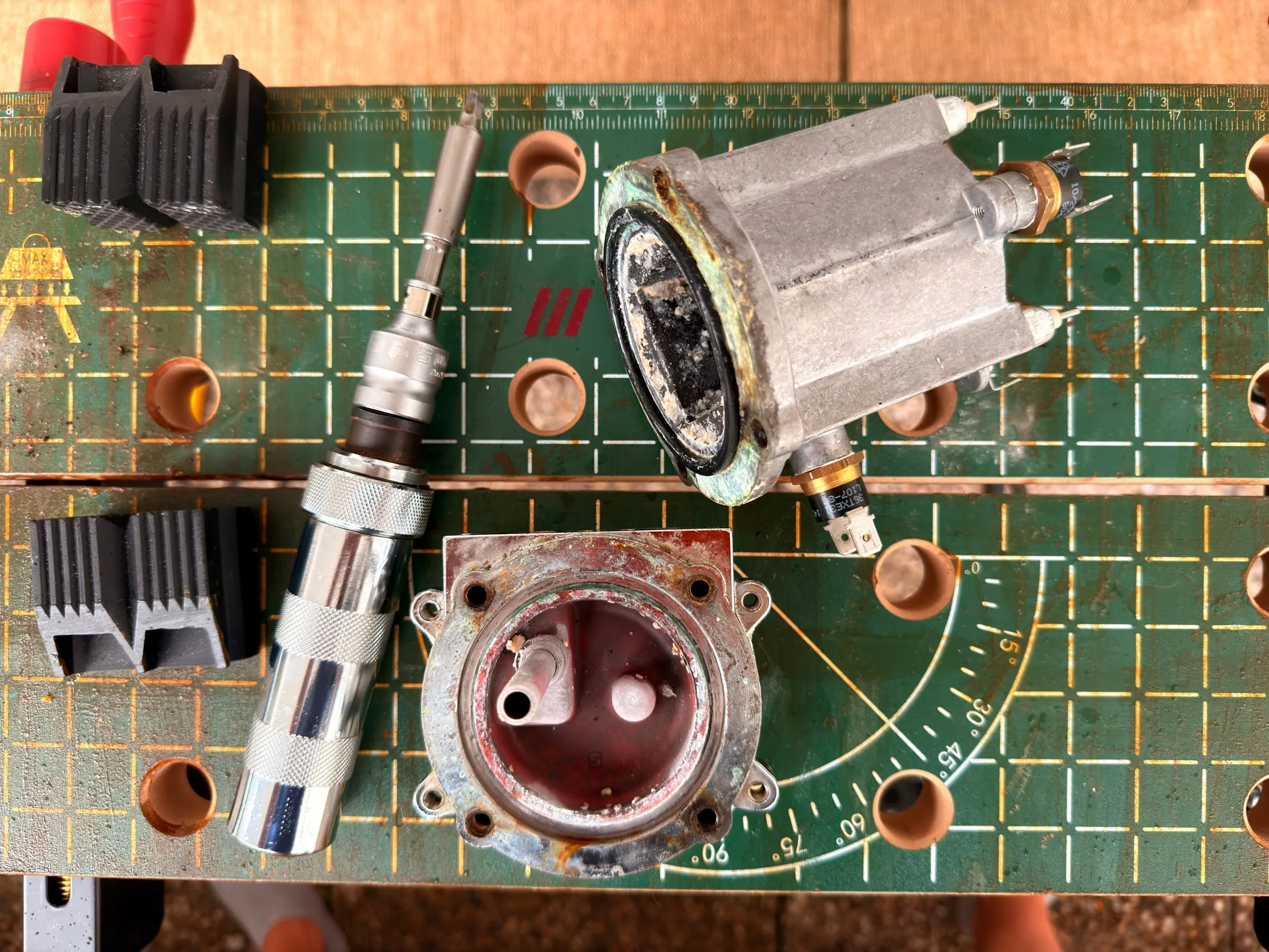 The opened boiler on a green workbench: the aluminium top half with the heating element lying on its side, the base with its red gasket in front, and the chrome impact driver that did the job between them
