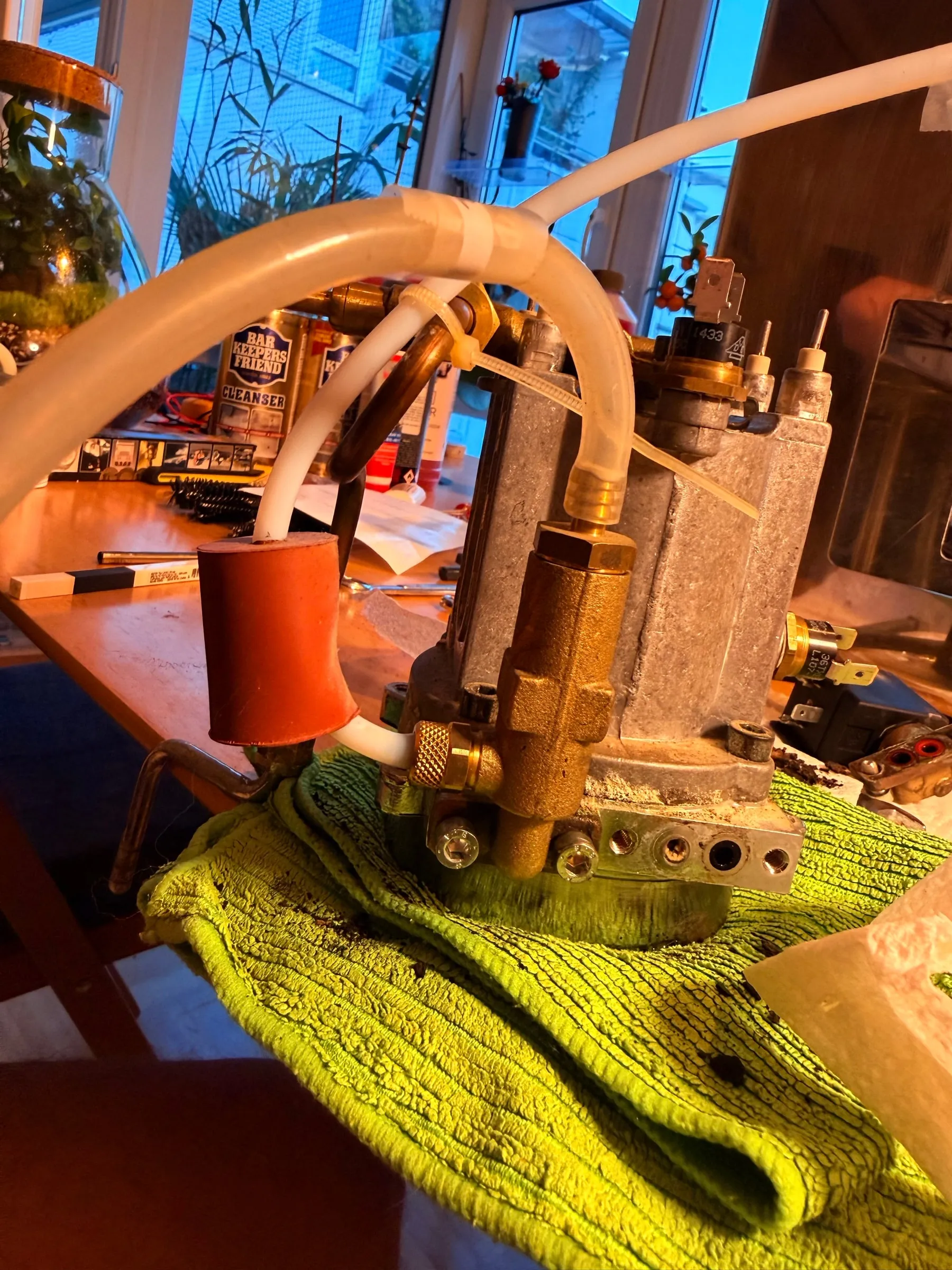 The removed boiler with the brew group standing on a green microfibre cloth, its aluminium flank white with limescale, silicone hoses looped over it and cleaning products in the background