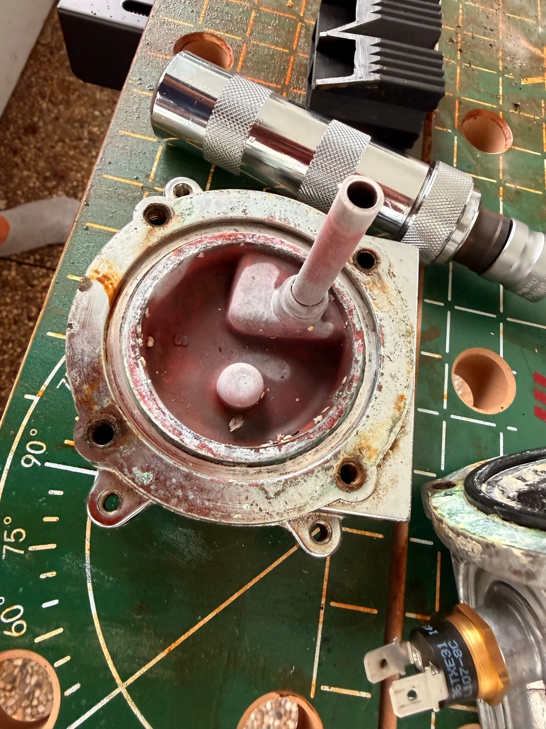 Close-up of the boiler base after opening: a dull red gasket ring, rust-brown deposits around the screw holes and the outlet tube, the knurled grip of the impact driver at the edge of the frame