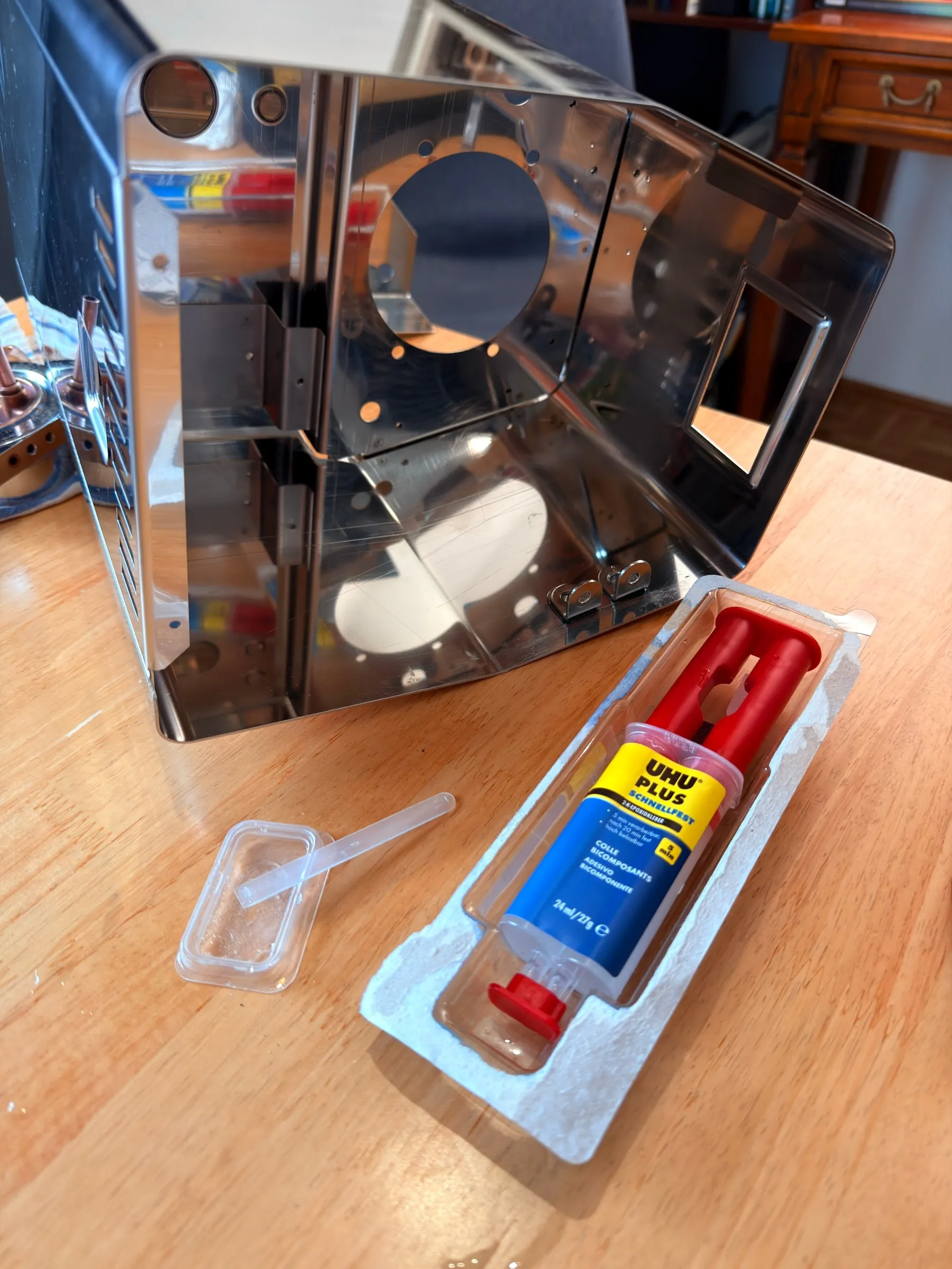 The polished stainless housing standing on the wooden table on the evening of 18 July, a pack of UHU Plus two-part epoxy with its mixing spatula lying in front of it