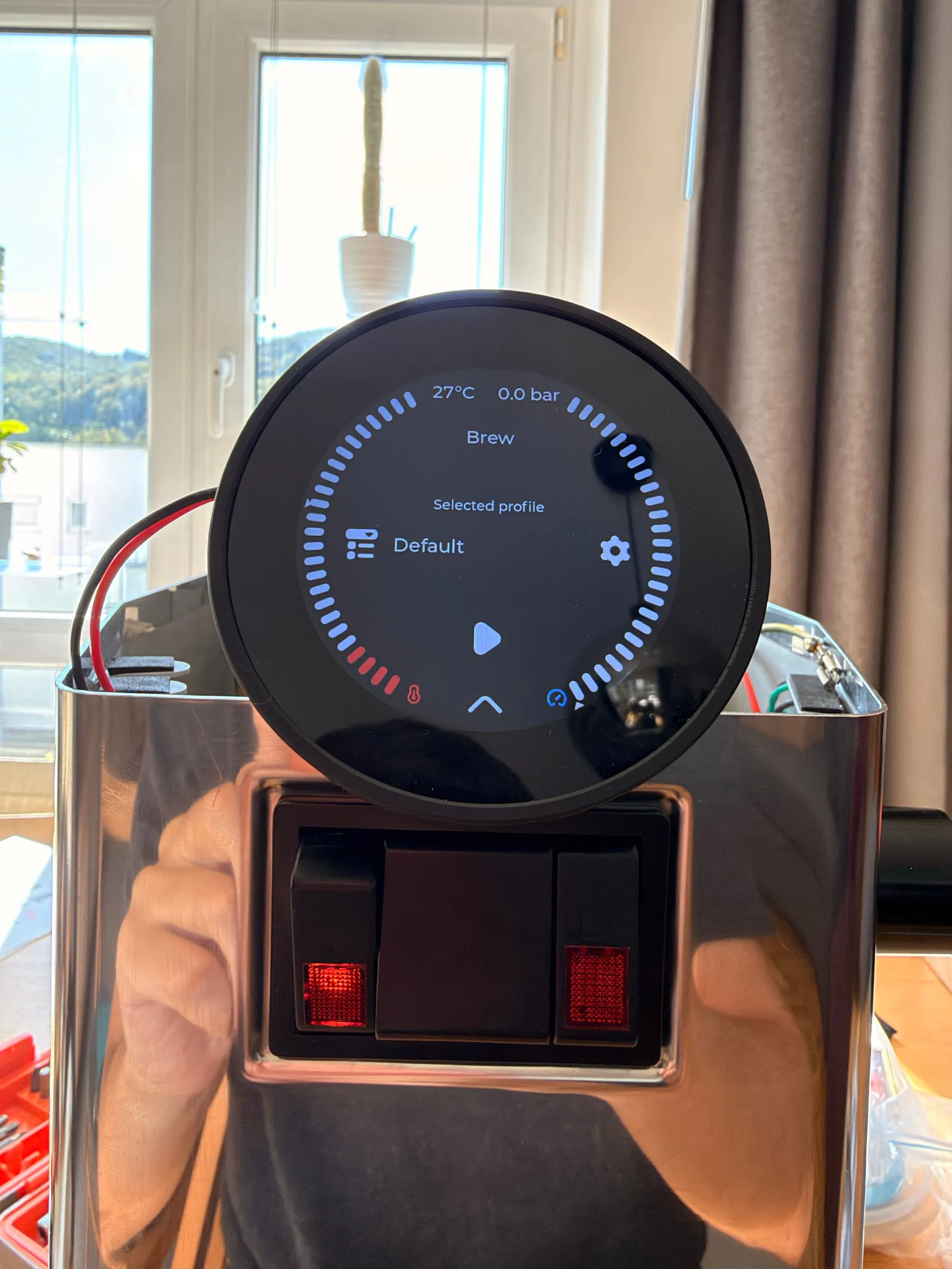 The round display on the machine in front of a window showing the brew screen with 27 degrees and 0.0 bar in the header and the profile Default