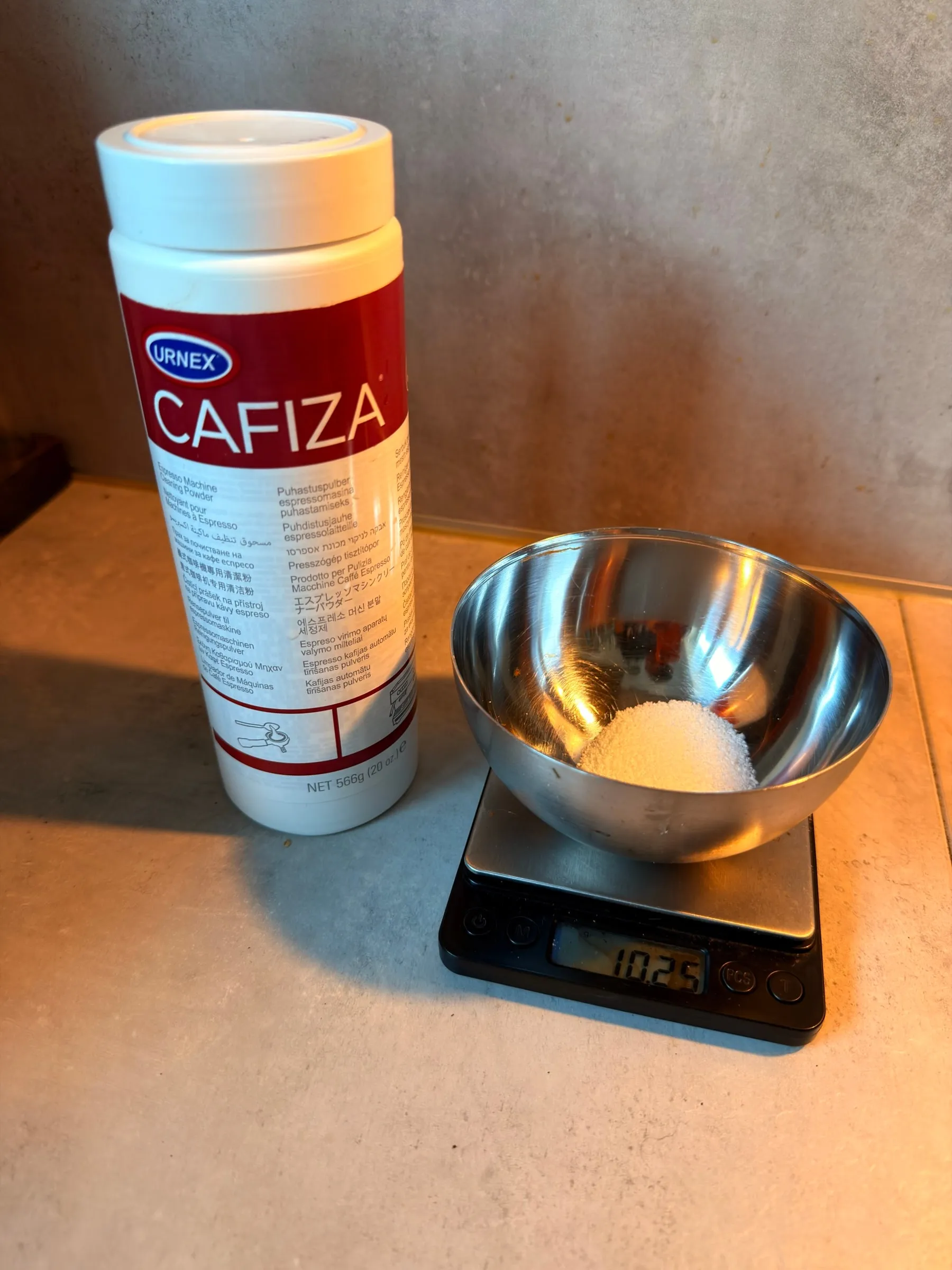 A tub of Urnex Cafiza next to a small steel bowl of the white powder on a pocket scale
