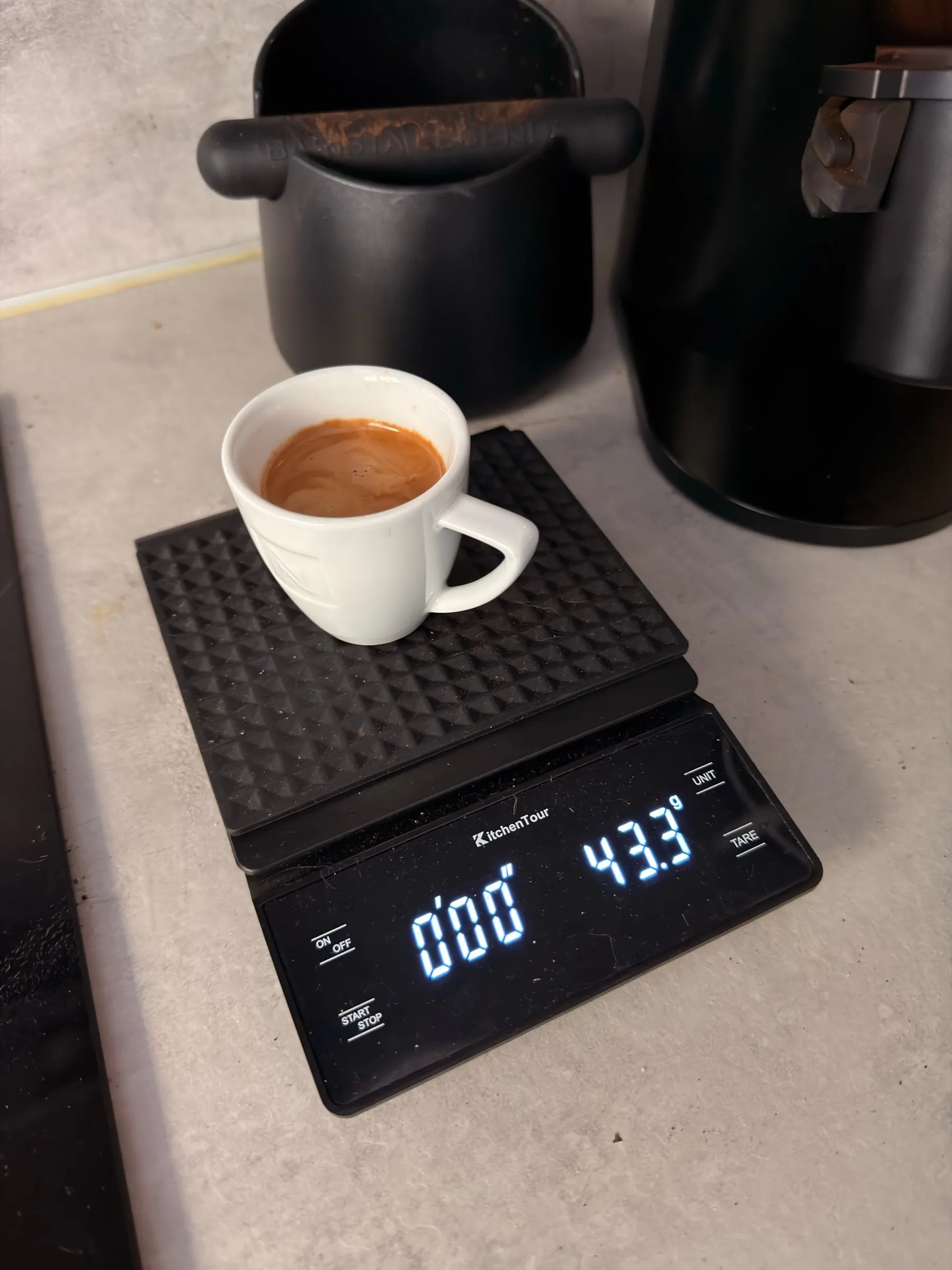 A small white espresso cup filled with crema standing on a black scale on the worktop, the scale reading 43.3 grams, a knock box behind it