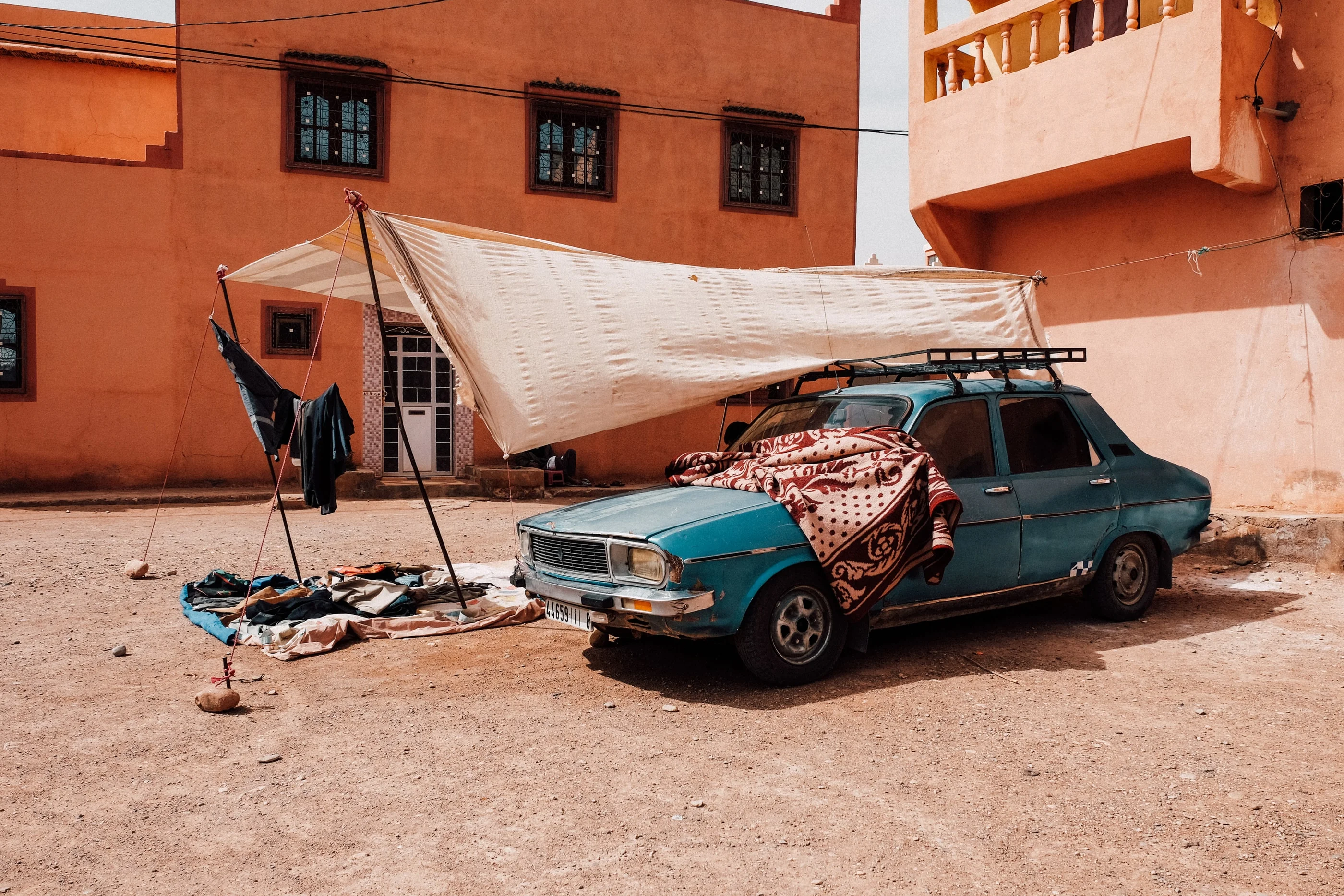 Old blue car under a tarpaulin in front of terracotta houses, a rug and laundry drying beside it