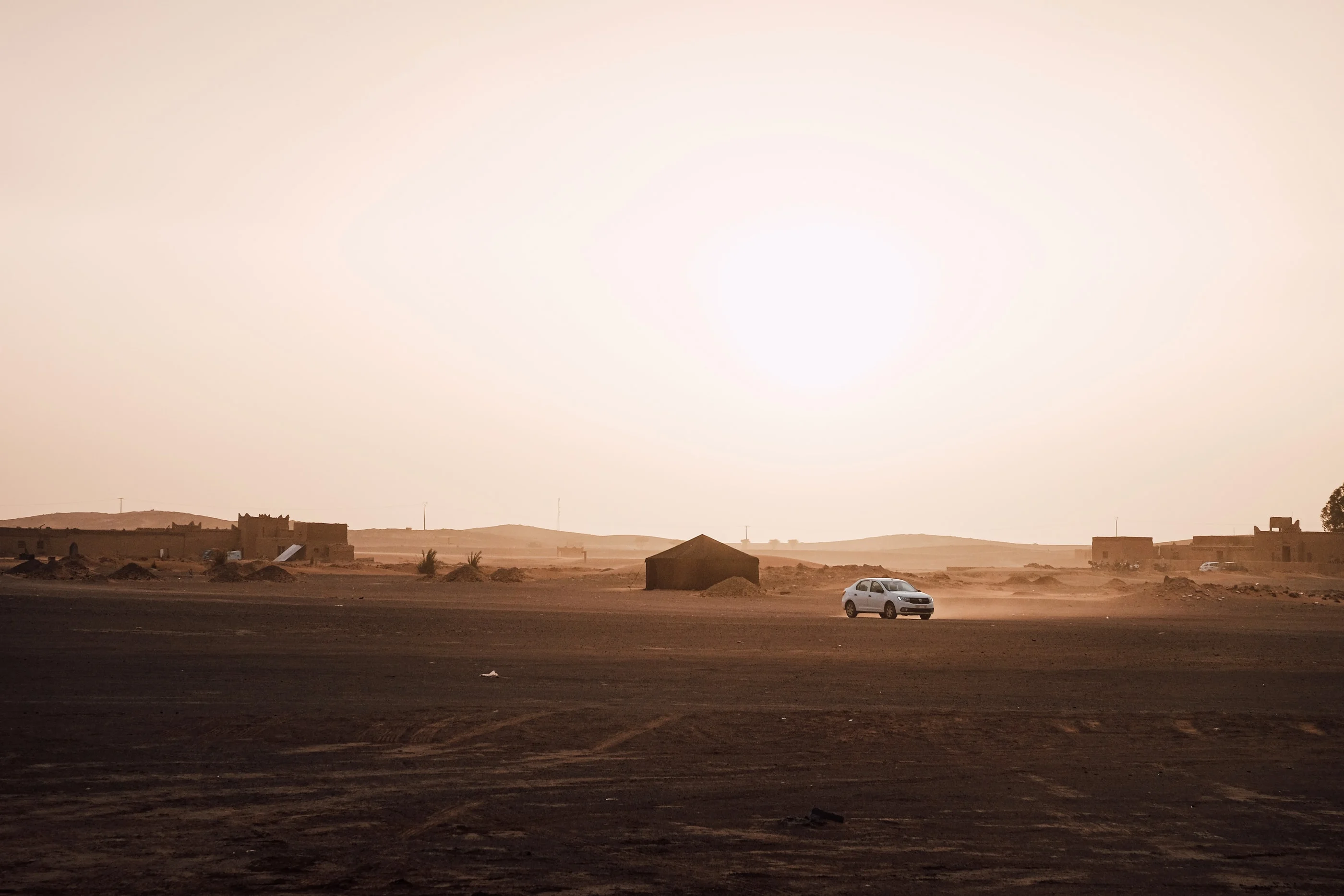 Off-road car on a dark desert plain at sunset, low buildings on the horizon