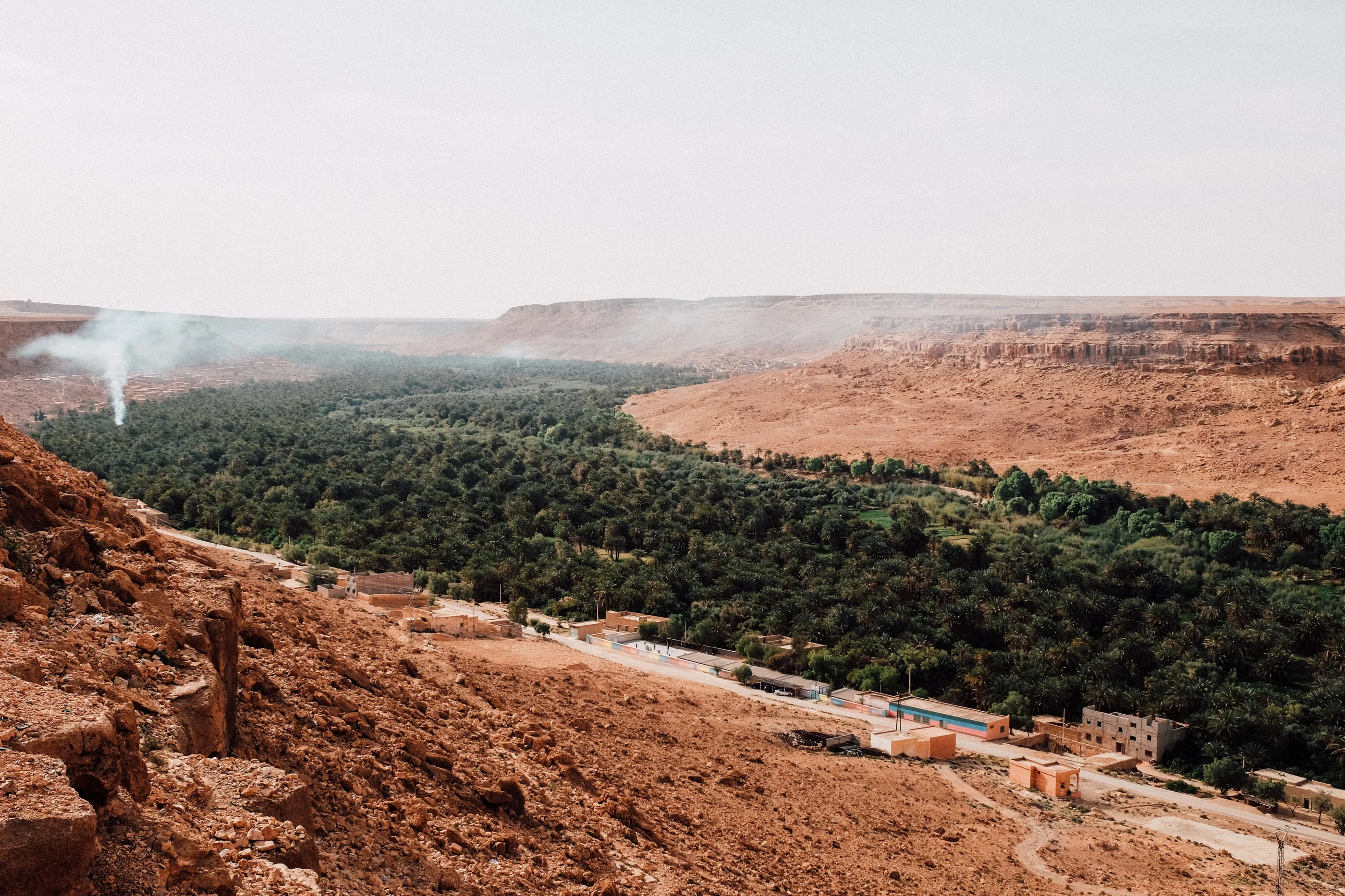 Long green palm oasis in a valley between red cliffs, smoke rising in the distance