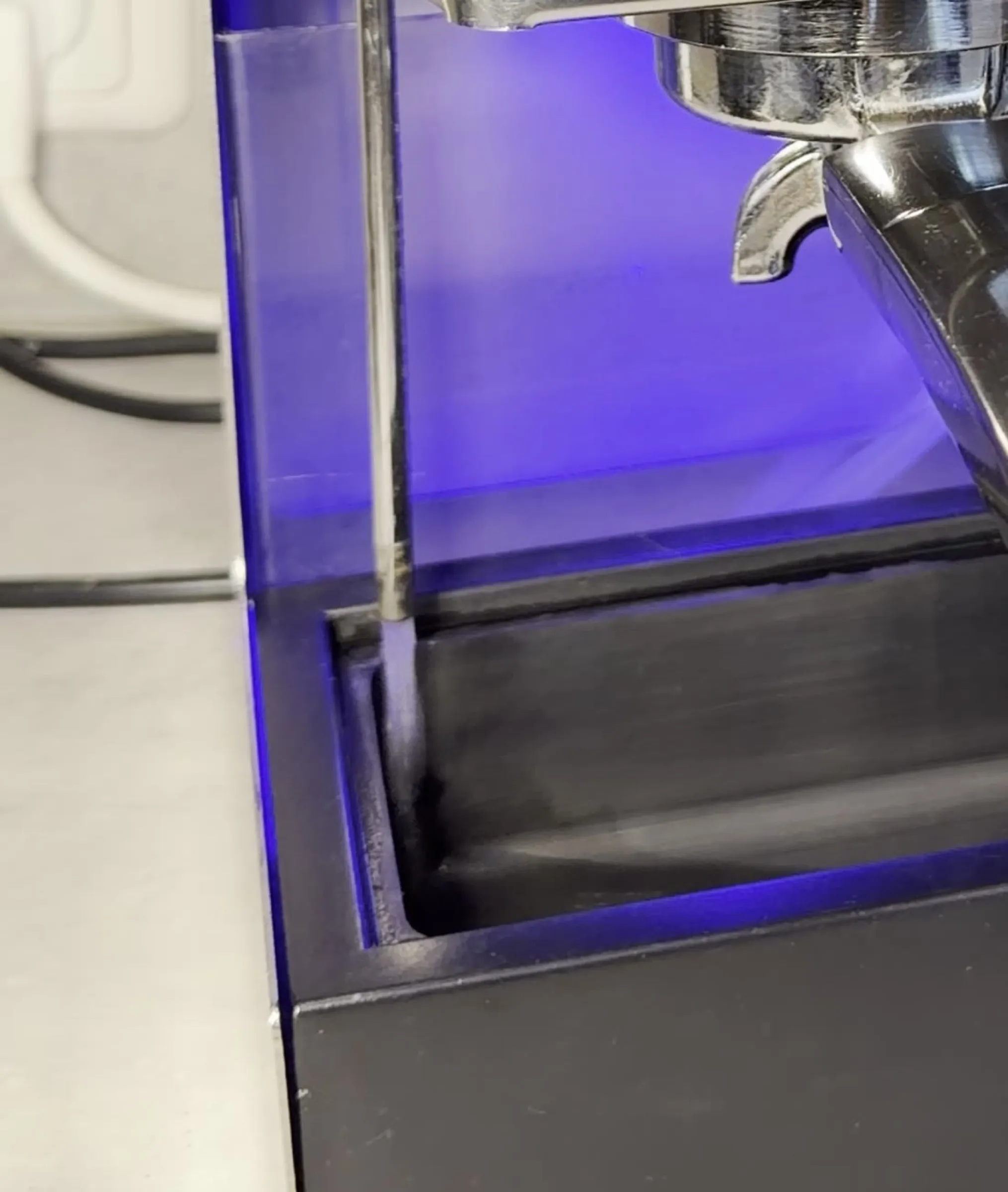 Close-up of the drain pipe of the solenoid valve above the drip tray with the grid removed, a thin jet of water running from the pipe, the tray lit blue