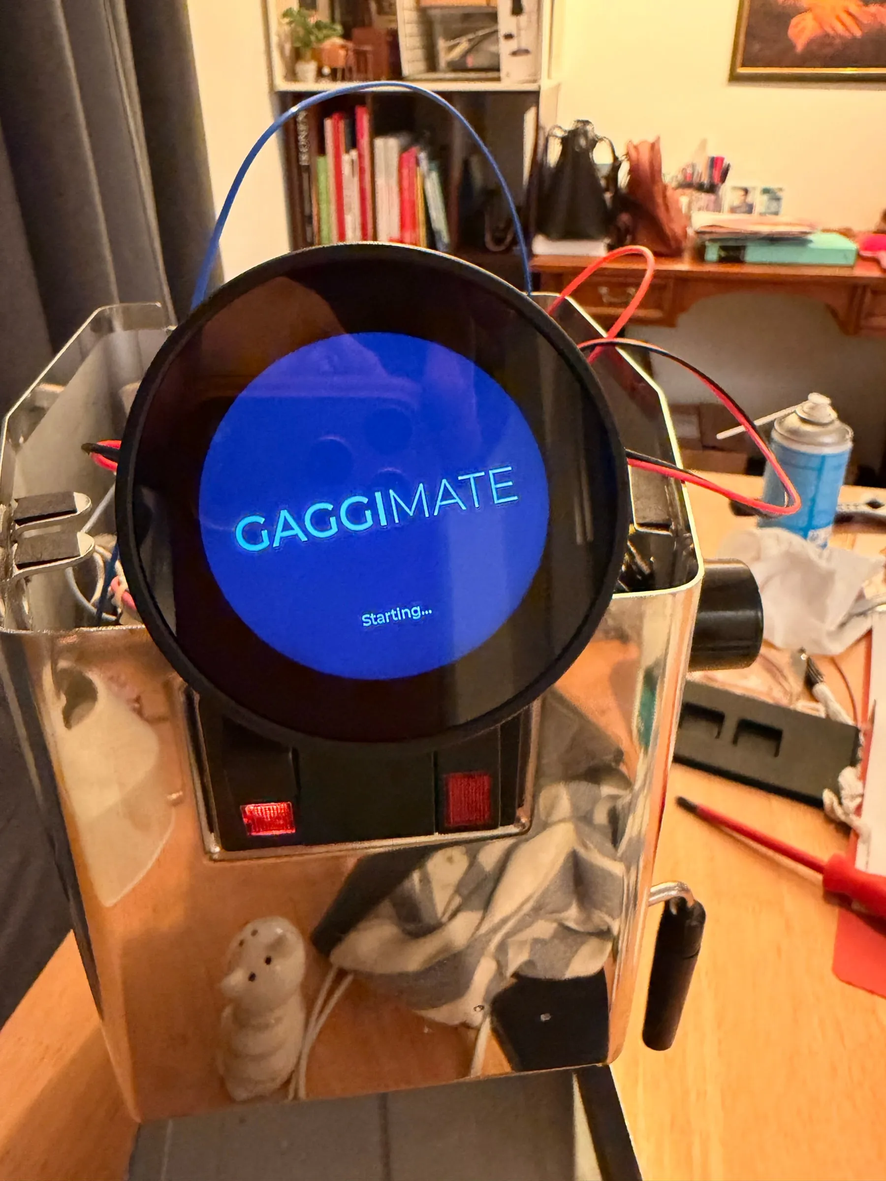 The round display mounted above the switch block of the open machine on the living room table, showing the GaggiMate logo on a blue background with the word Starting below it