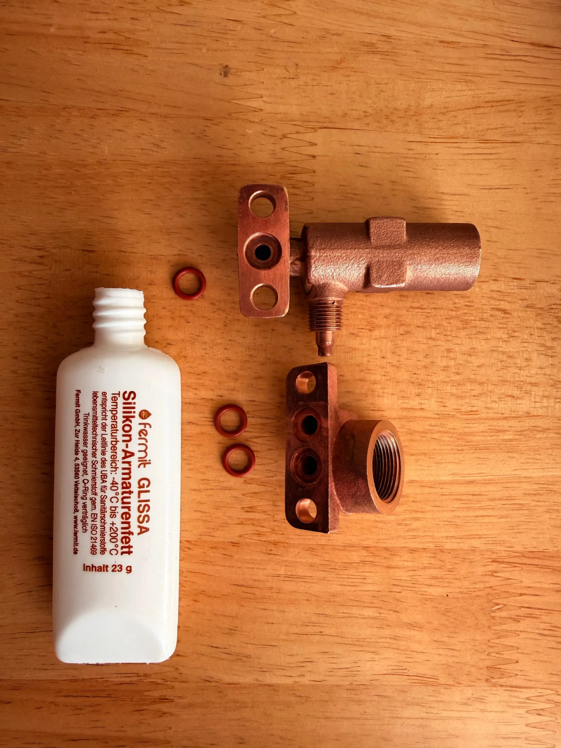 A white tube of Fermit Glissa silicone grease on a wooden table next to the cleaned brass over-pressure valve, the solenoid valve body and four small red flange gaskets