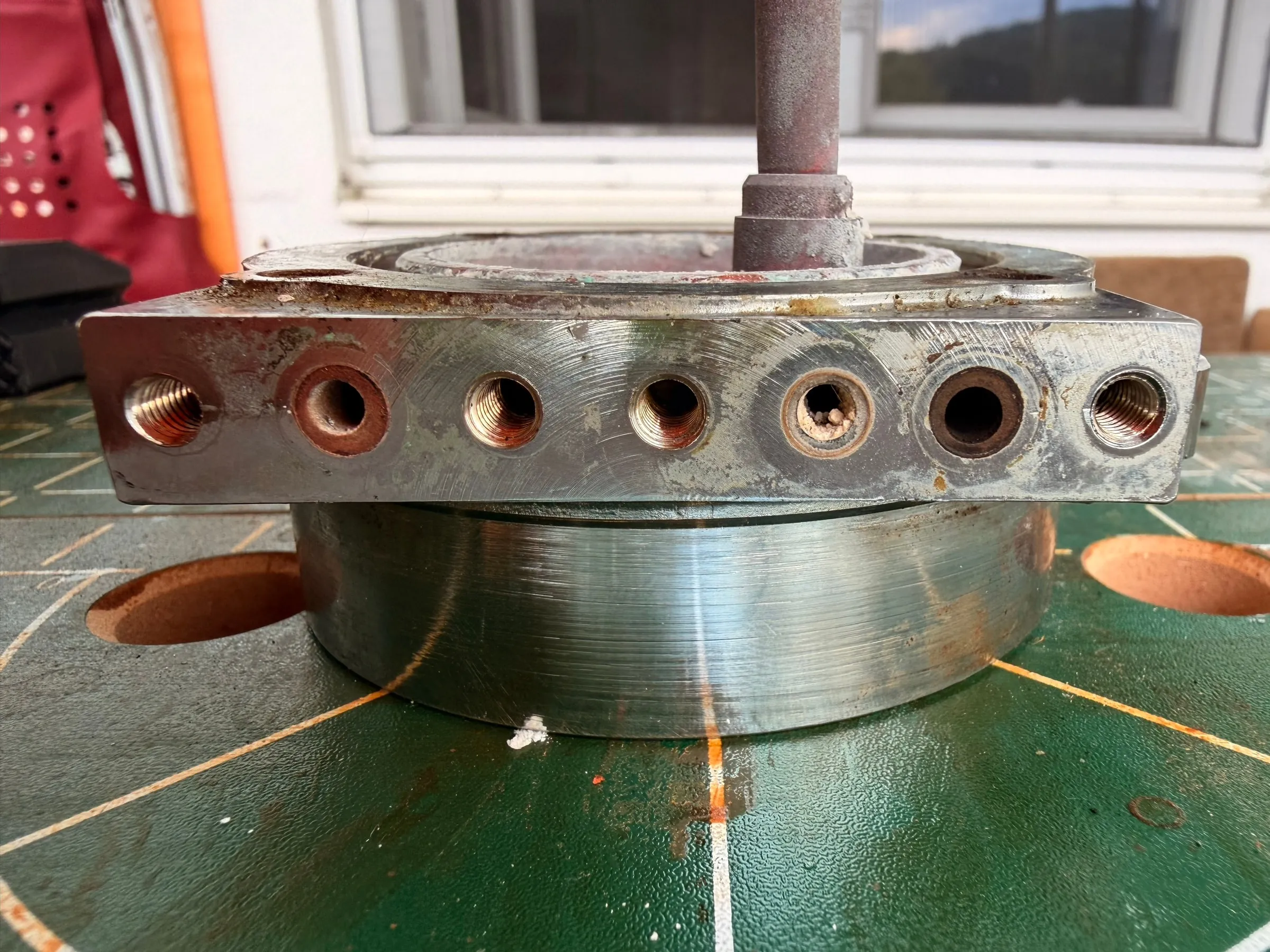 The group body on 18 July, seen from the side on a green cutting mat with the copper standpipe rising from it: the machined sealing face is coated with white scale, rust-brown deposits sit around the threaded holes of the flange