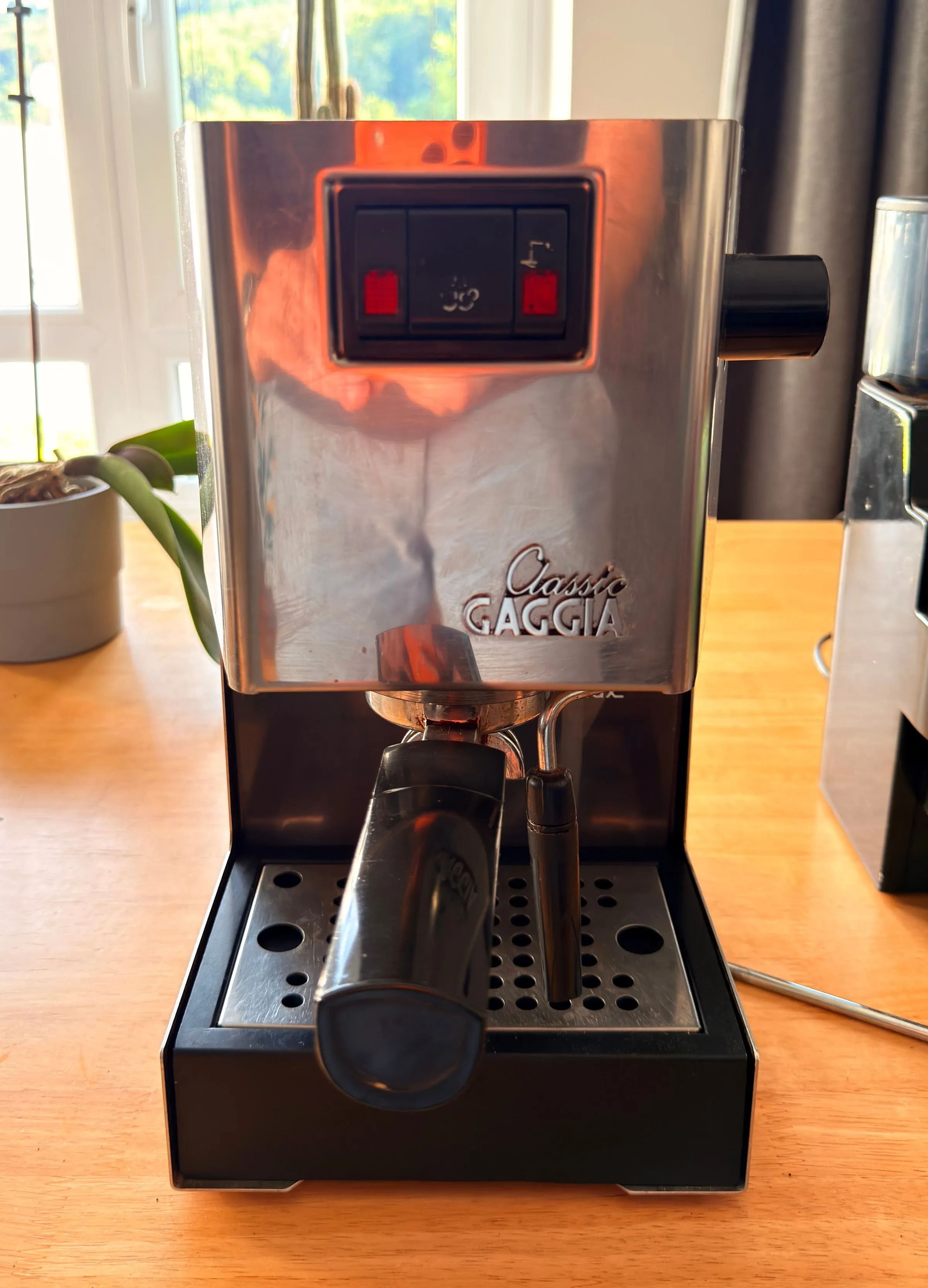 A Gaggia Classic espresso machine on a wooden kitchen table in warm evening light, seen from the front with the portafilter locked in, its polished steel reflecting the room, the edge of a Gaggia grinder beside it
