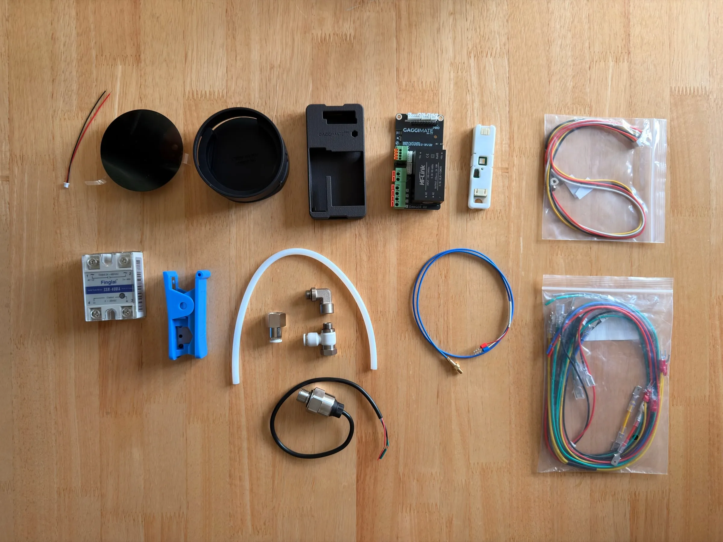 The GaggiMate Pro kit laid out on a wooden table: two round display parts, a black printed housing with the green control board inside, a small module in a bag, the white solid-state relay, a blue clip, a length of white PTFE hose with brass and plastic fittings, the black pressure sensor on its cable, a blue probe cable and two bags of pre-made cable sets
