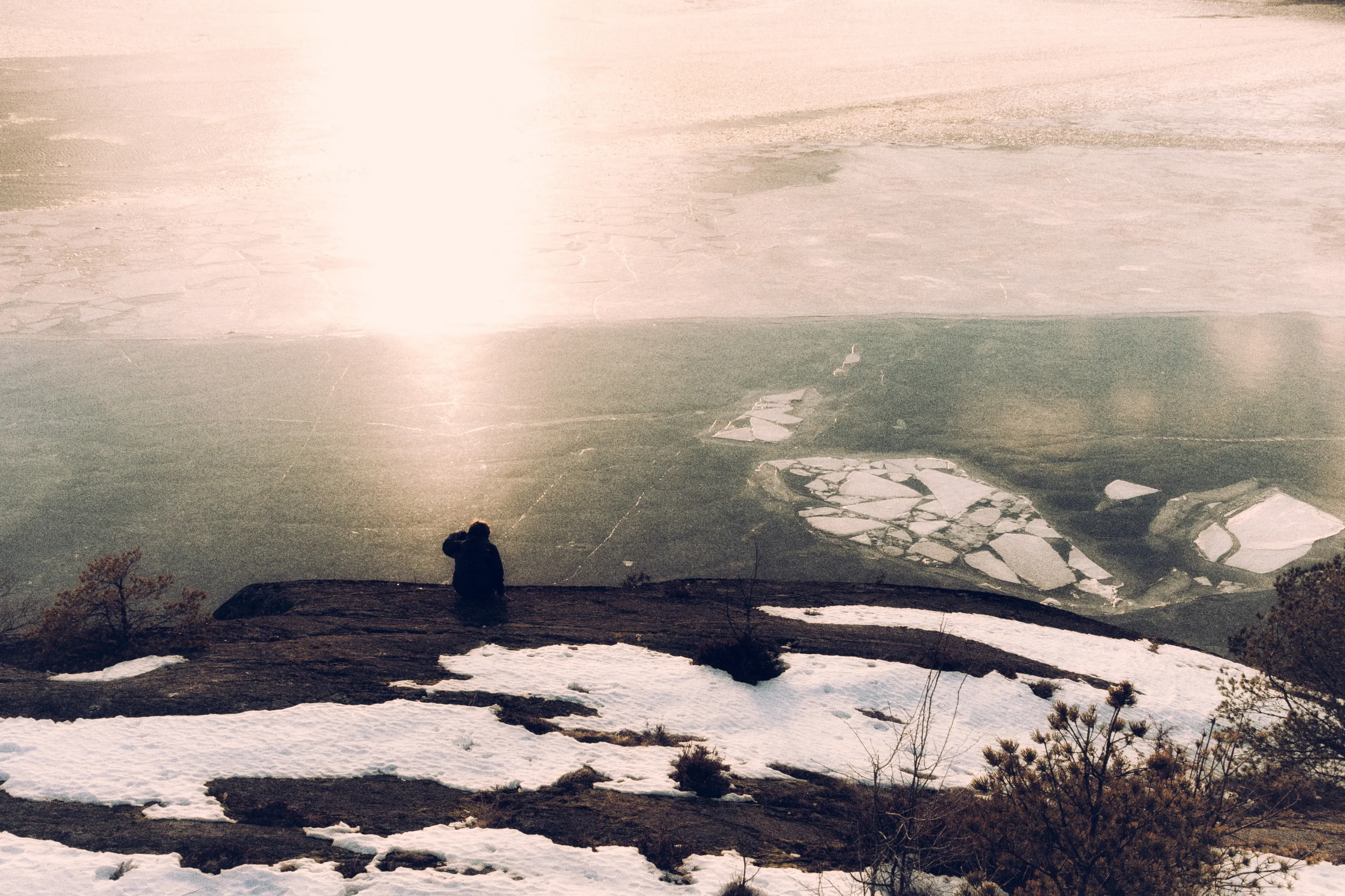 Silhouette of a person sitting on a snowy rock above a frozen lake, the low sun reflecting on the ice