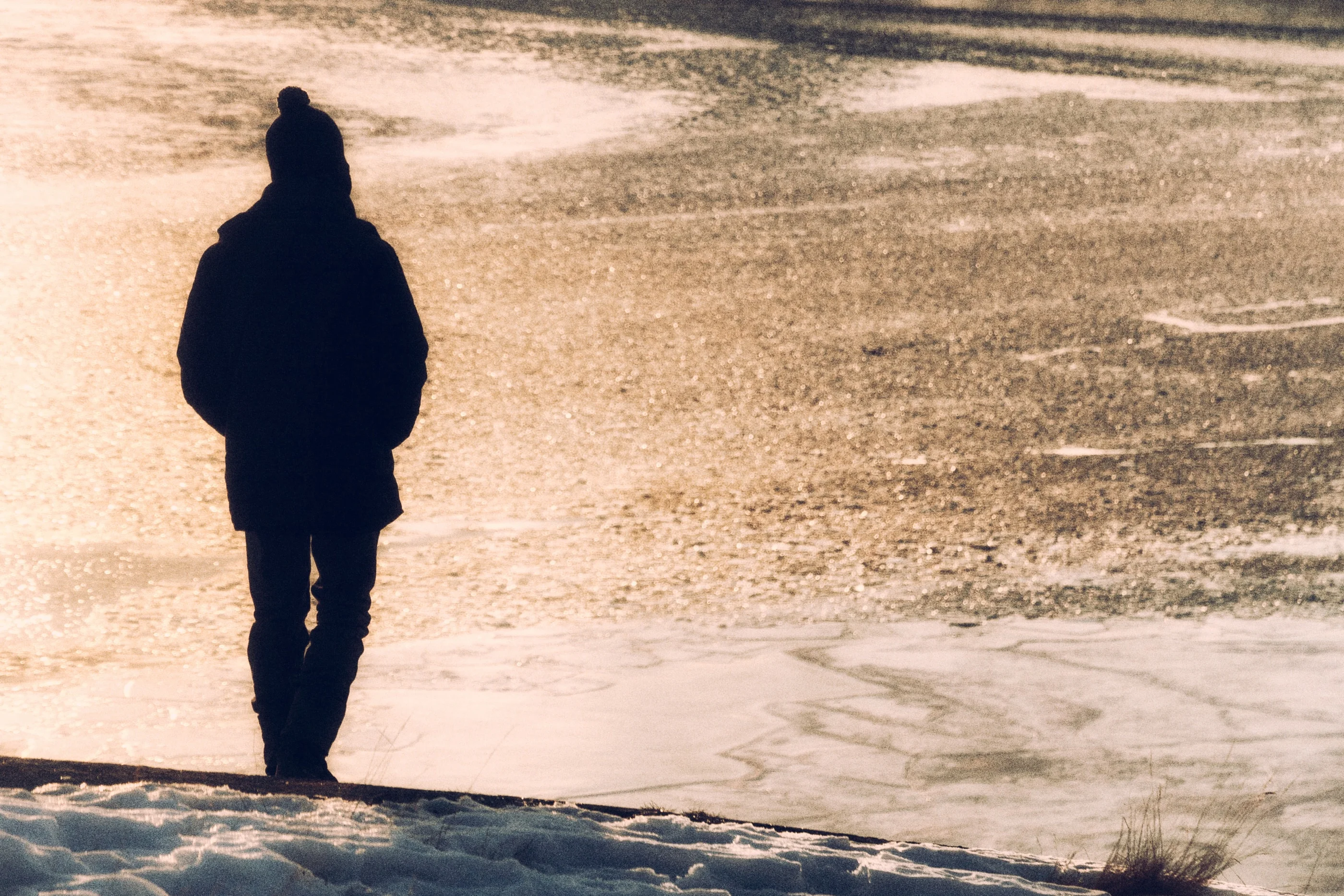 Silhouette of a person in a beanie standing on the shore of an icy lake