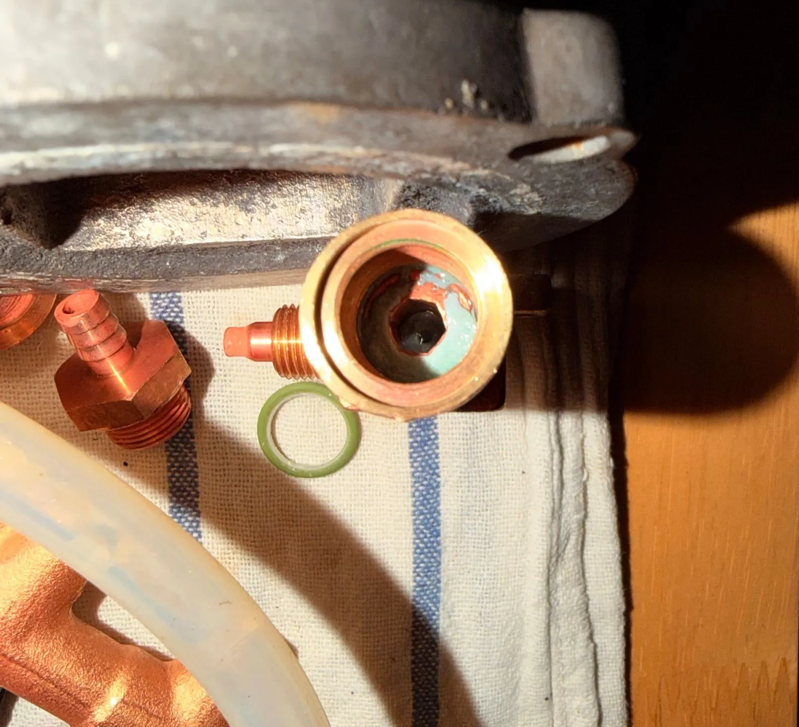 Looking into the brass over-pressure valve under the boiler flange: the internal hex adjuster sits deep in the bore, ringed with green verdigris, a green O-ring and brass fittings lying on a tea towel beside it