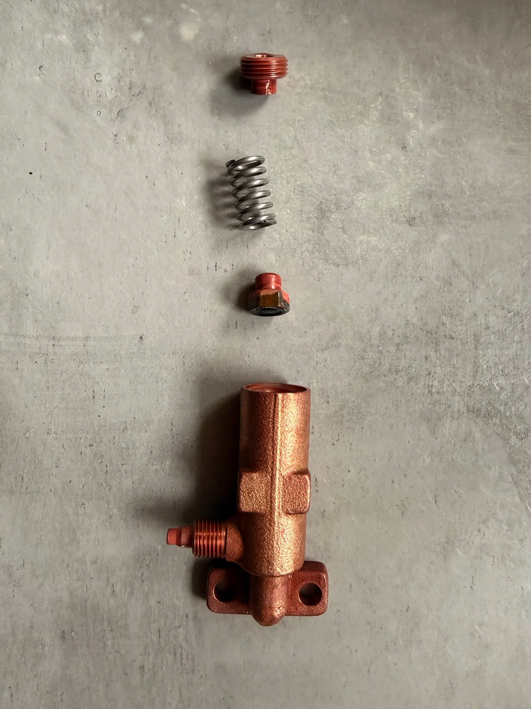 The over-pressure valve laid out in disassembly order on a grey surface: the small threaded adjuster at the top, then the spring, the piston and the brass valve body with its flange