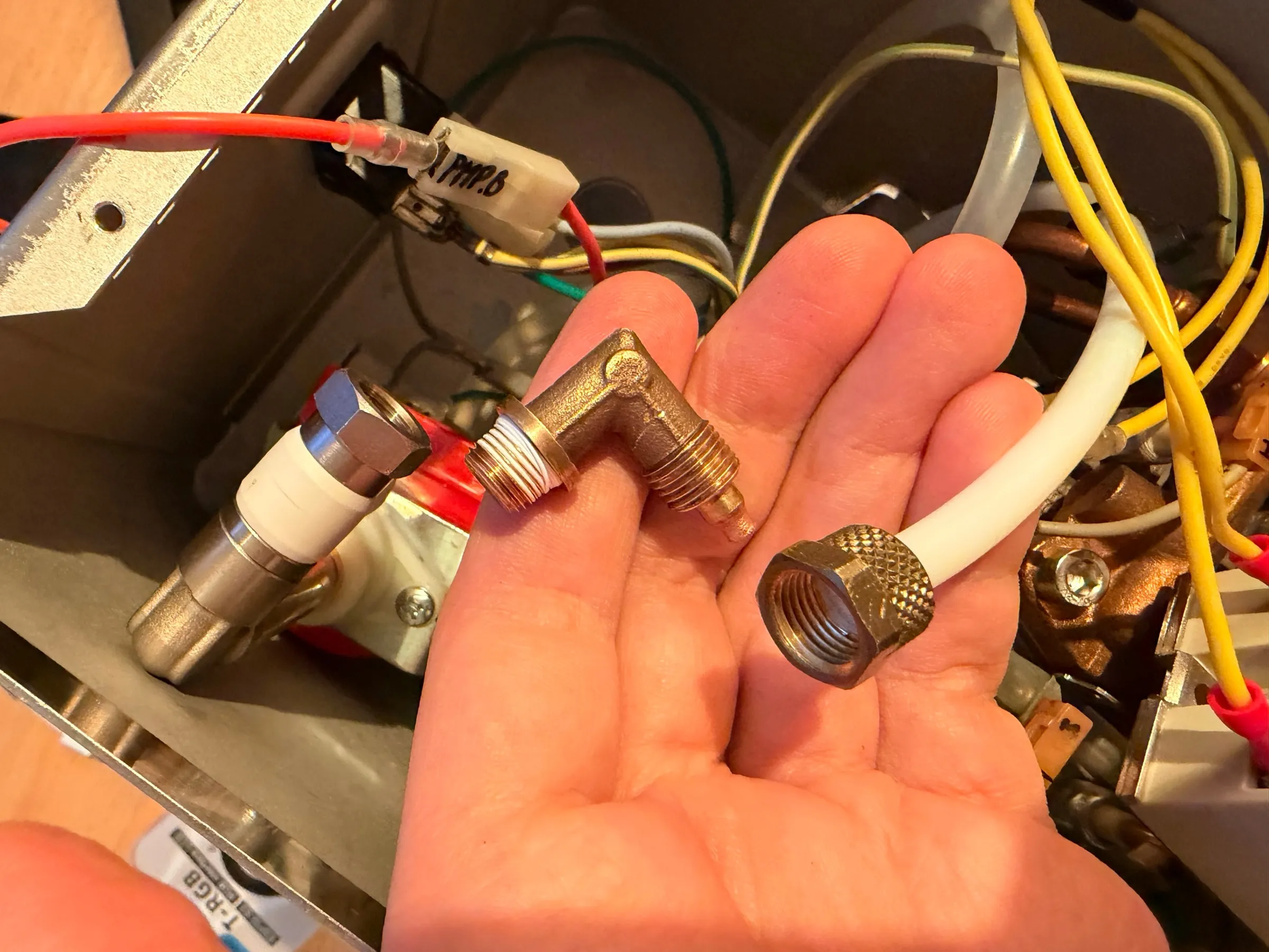 A hand holding the brass elbow fitting removed from the pump and the knurled nut with the white PTFE hose, the labelled wires and yellow relay cables in the open housing behind