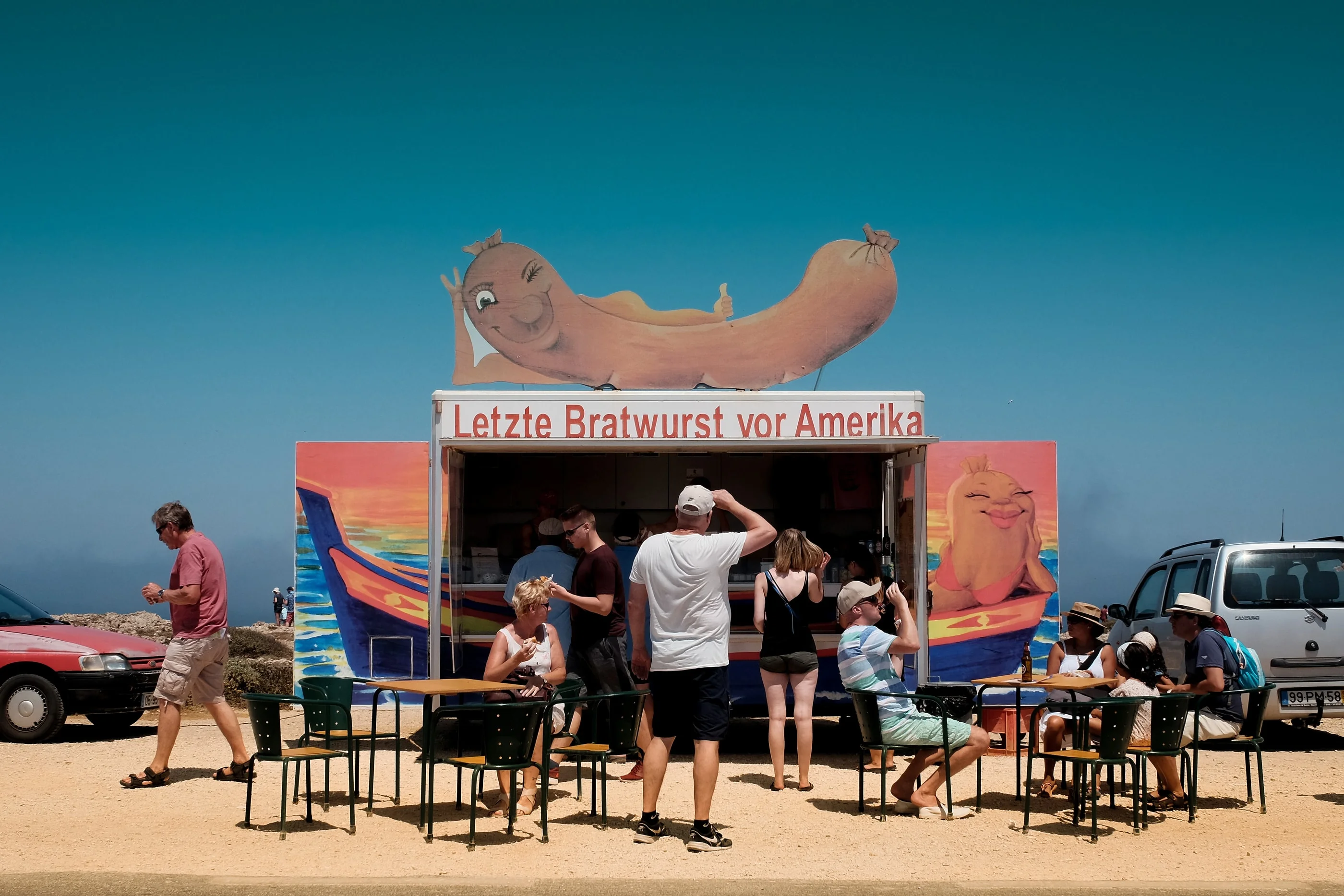 Snack bar with a giant bratwurst sign reading 'Letzte Bratwurst vor Amerika', visitors at the tables