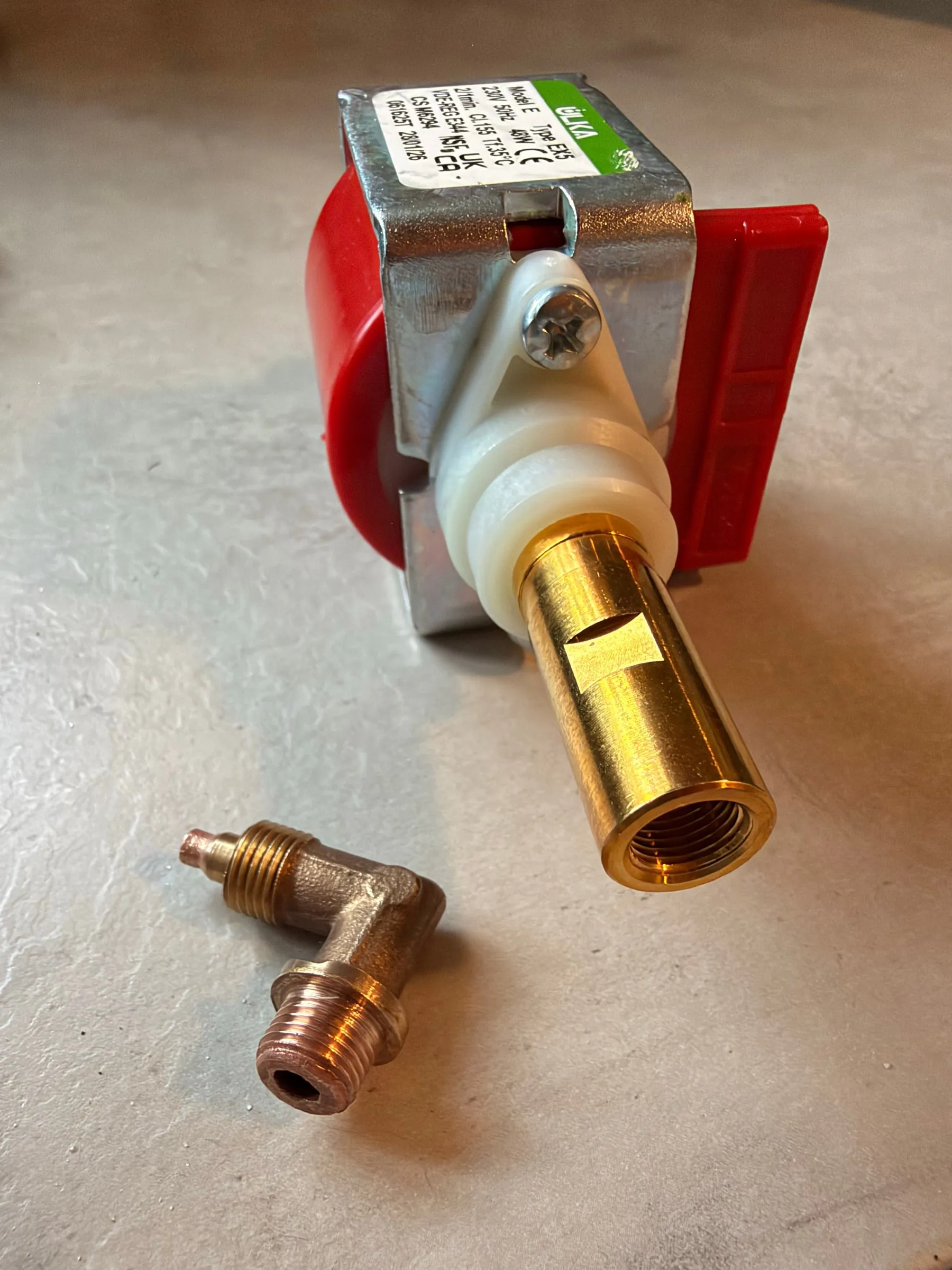 The new Ulka EX5 pump lying on a grey surface, its green specification label legible and the brass outlet collar prominent, the brass elbow fitting in the foreground