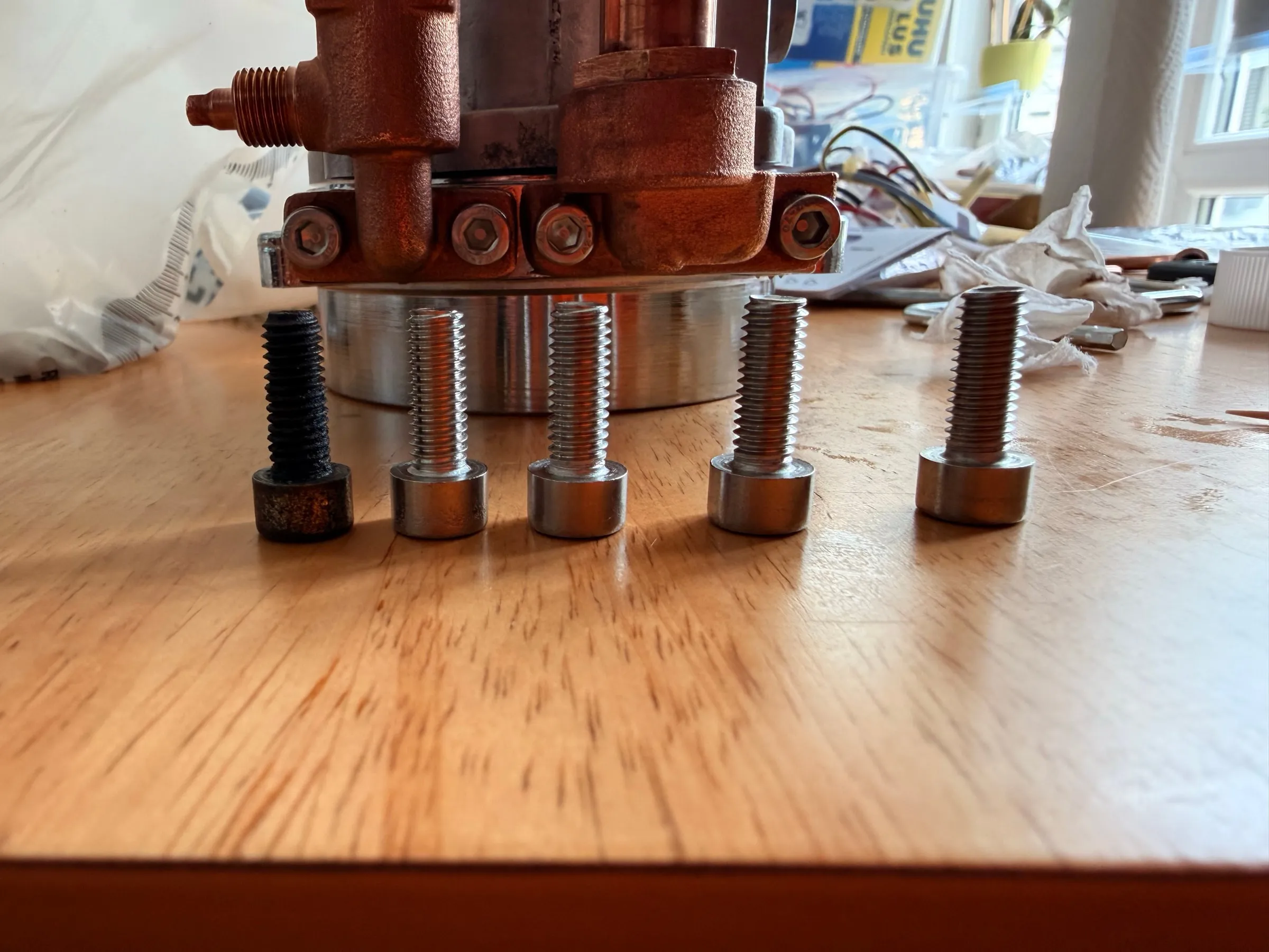 One black rust-pitted original screw standing next to four bright new stainless hex-socket screws, the freshly bolted boiler behind them