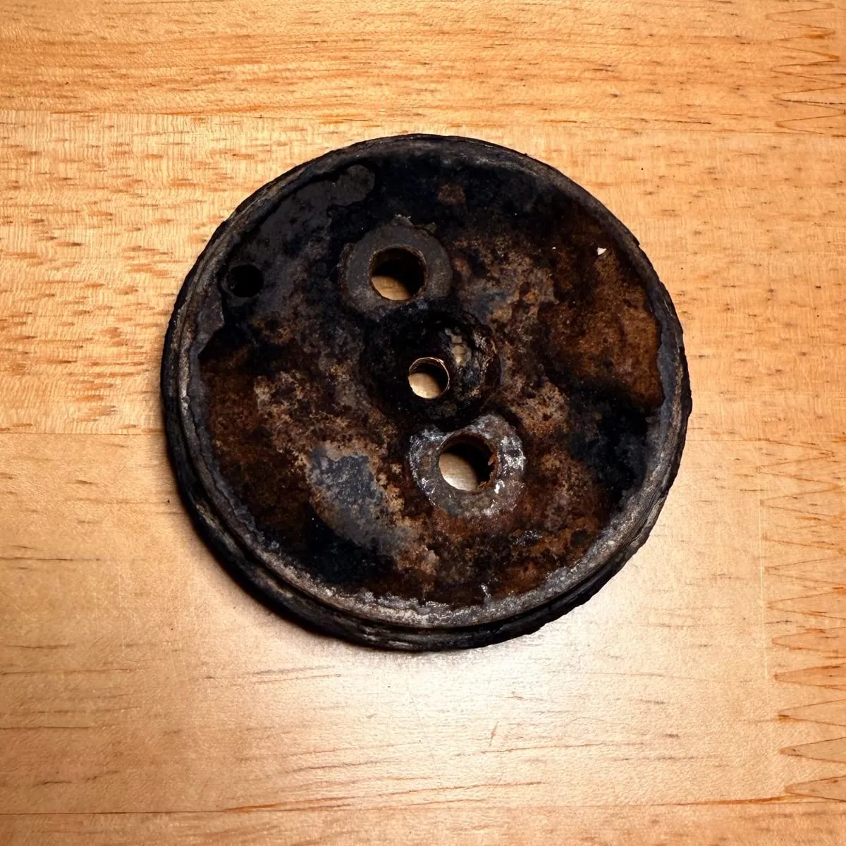 The old aluminium shower holder plate on a wooden table, coffee side up: a round disc with three holes, covered in a black and brown crust of old coffee