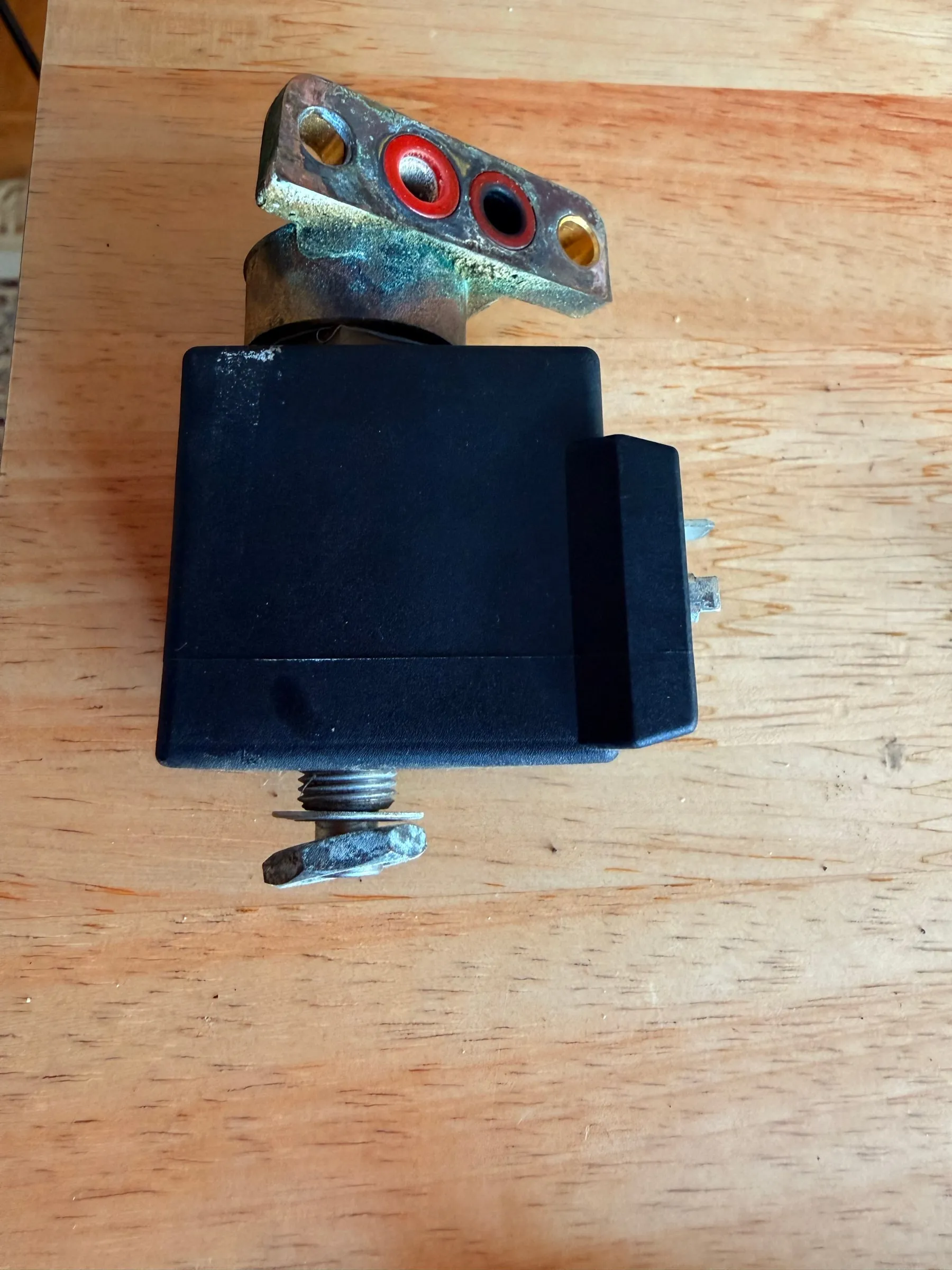 The solenoid valve with its black coil standing on a wooden table before cleaning: the brass flange with two ports and gasket remnants on top, green verdigris where the body meets the coil
