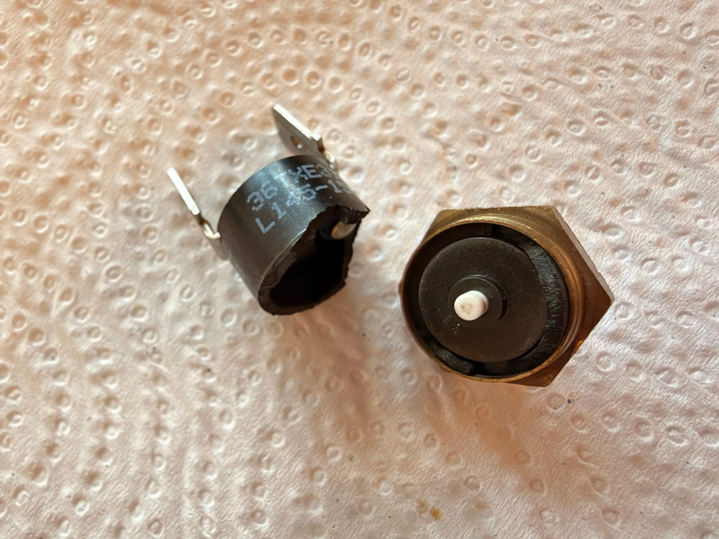 The broken steam thermostat on kitchen paper: the black plastic body with its two spade terminals lying on its side, and beside it the brass base with a white ceramic pin in its centre