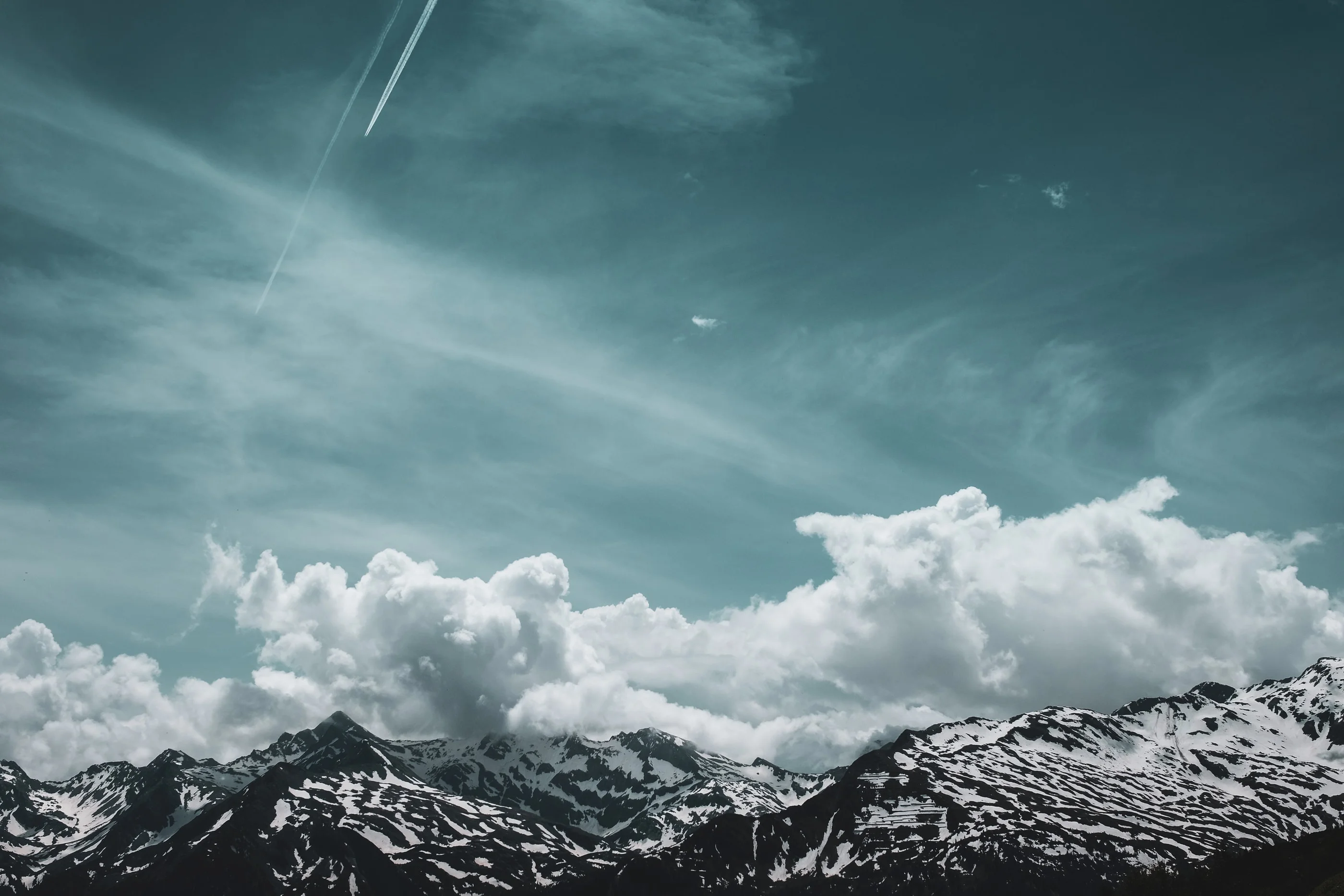 Snow-capped mountain range under clouds and a teal sky crossed by a contrail