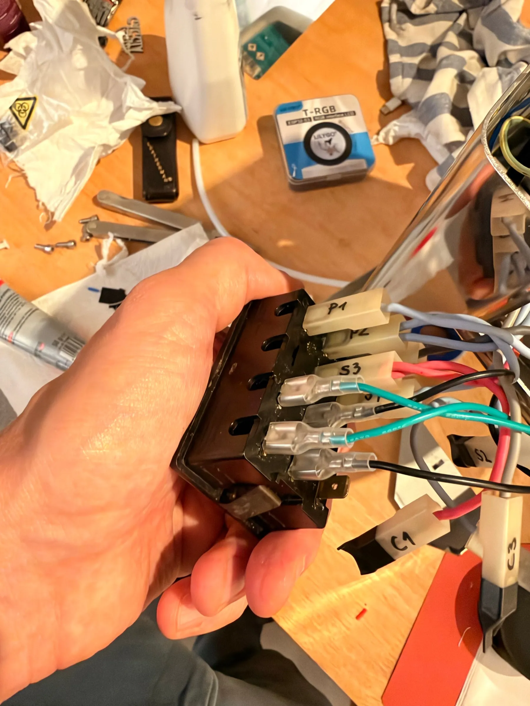 A hand holding the four-way switch block out of the front of the housing, the labelled wires P4, S3 and others plugged onto its spade terminals, the printed guide and cleaning products on the table behind