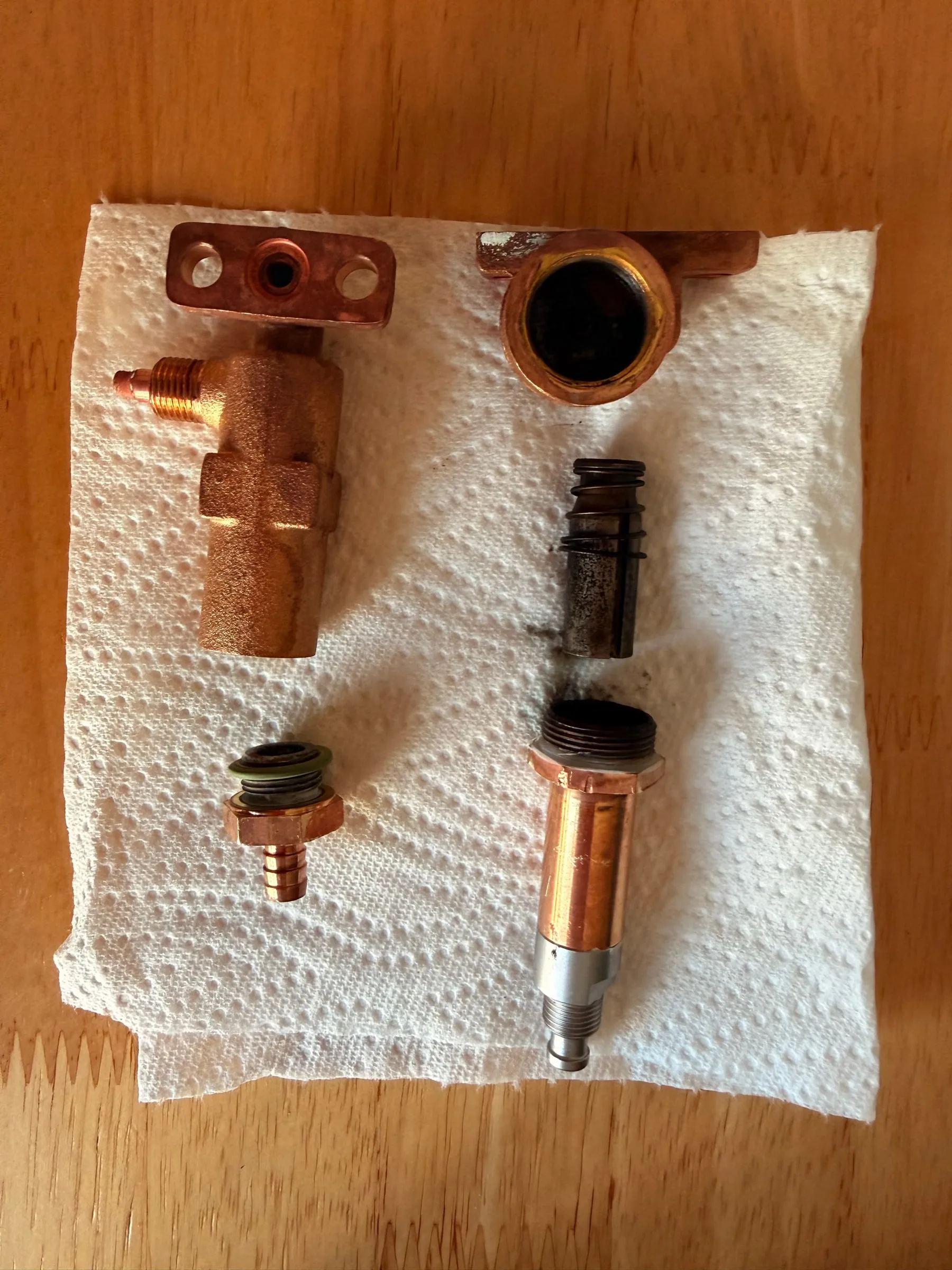 Valve parts laid out on kitchen paper. Left: the brass over-pressure valve body with its flange and a hose fitting with a green O-ring. Right: the solenoid valve body, its dark steel plunger with the spring, and the guide tube the plunger moves in