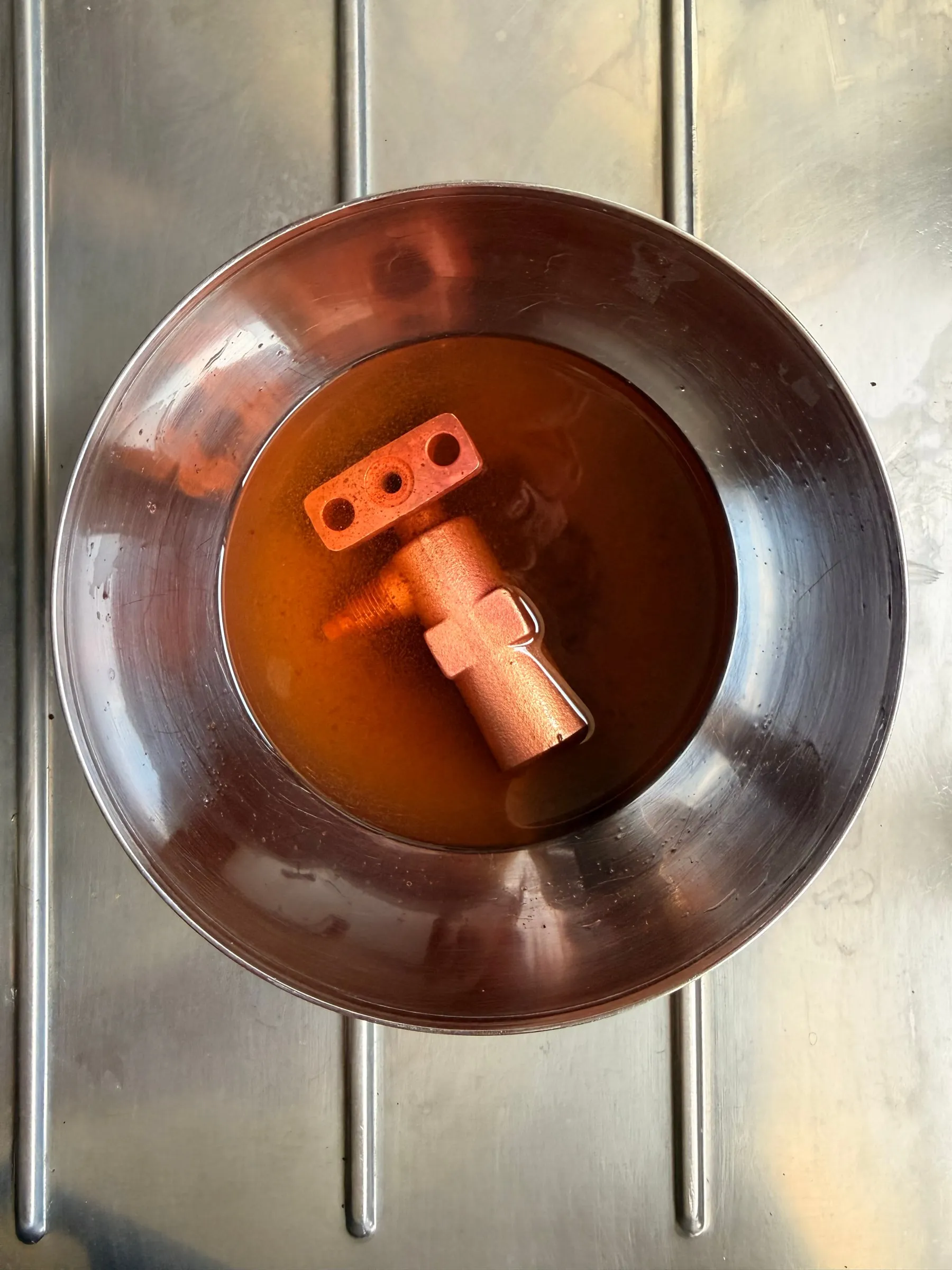 Looking down into a stainless bowl of amber liquid on a stove, a brass steam valve and a flange plate submerged and gently fizzing
