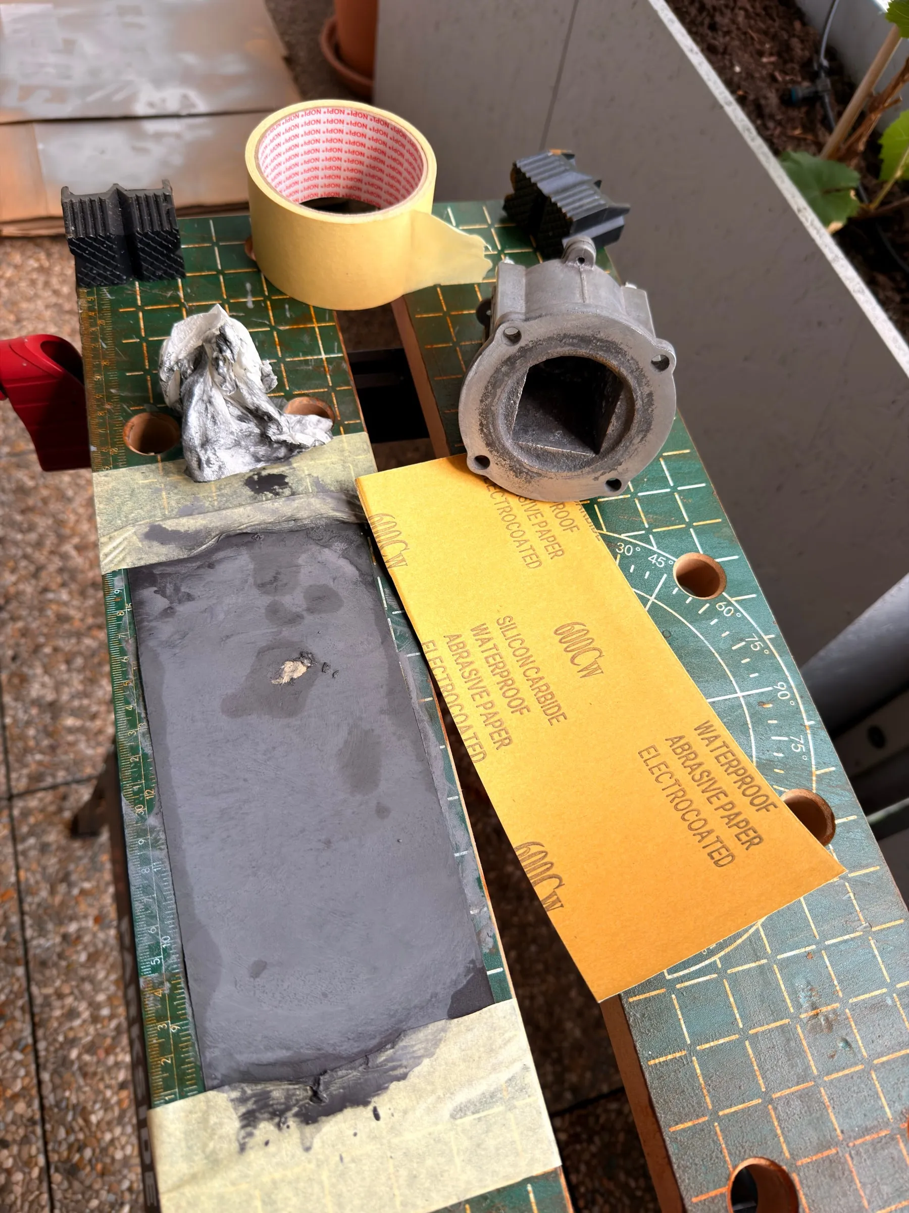 Wet-sanding setup outdoors on a green cutting mat: a strip of used grey sanding paper taped flat, a fresh sheet of 600 grit silicon-carbide paper, a roll of masking tape and the aluminium boiler part standing beside it