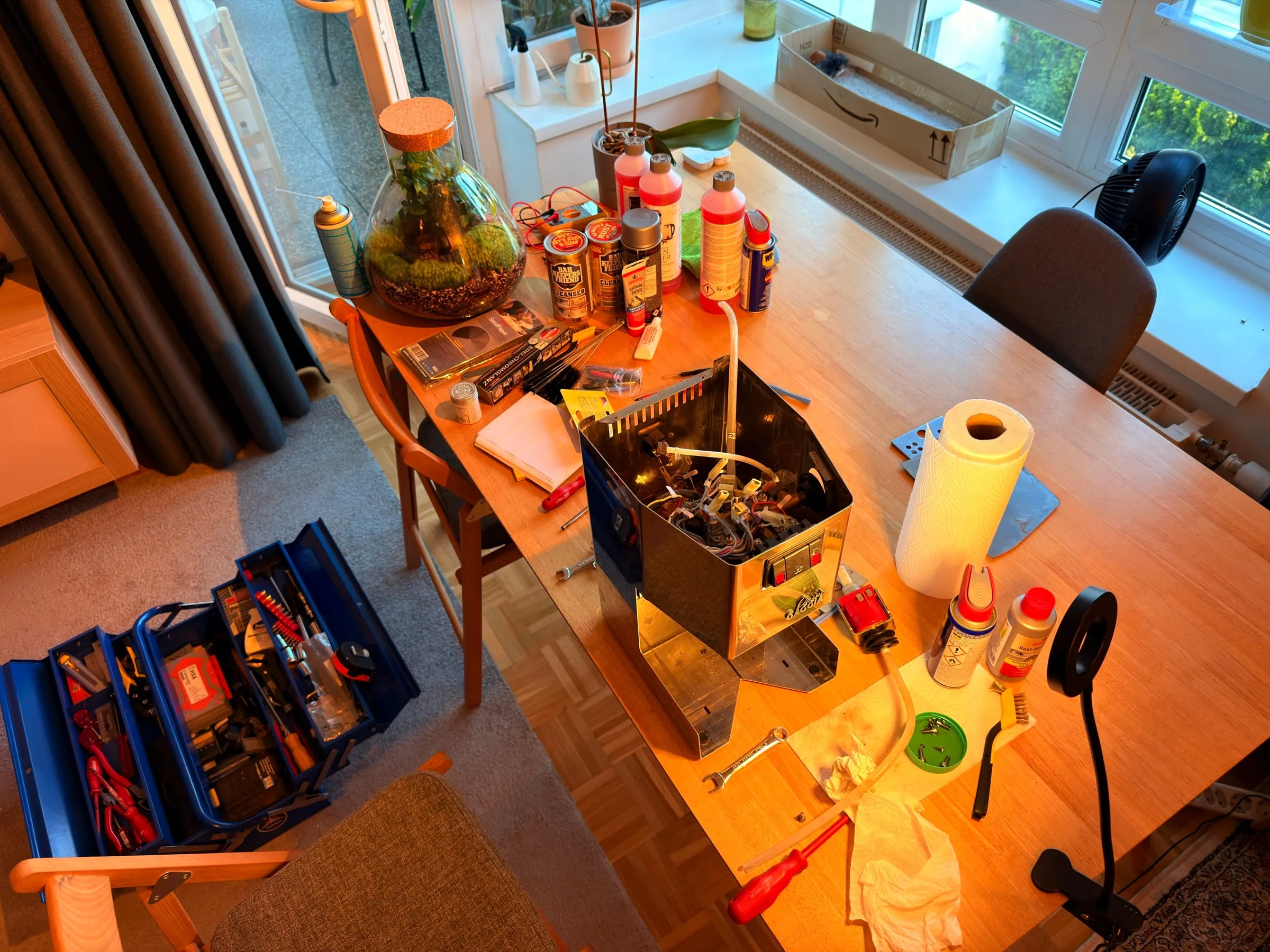 A living room table seen from above during the teardown: the opened housing full of loose cables in the middle, bottles and spray cans lined up at the back, kitchen paper, screwdrivers and a desk lamp around it, an open blue metal toolbox on the floor